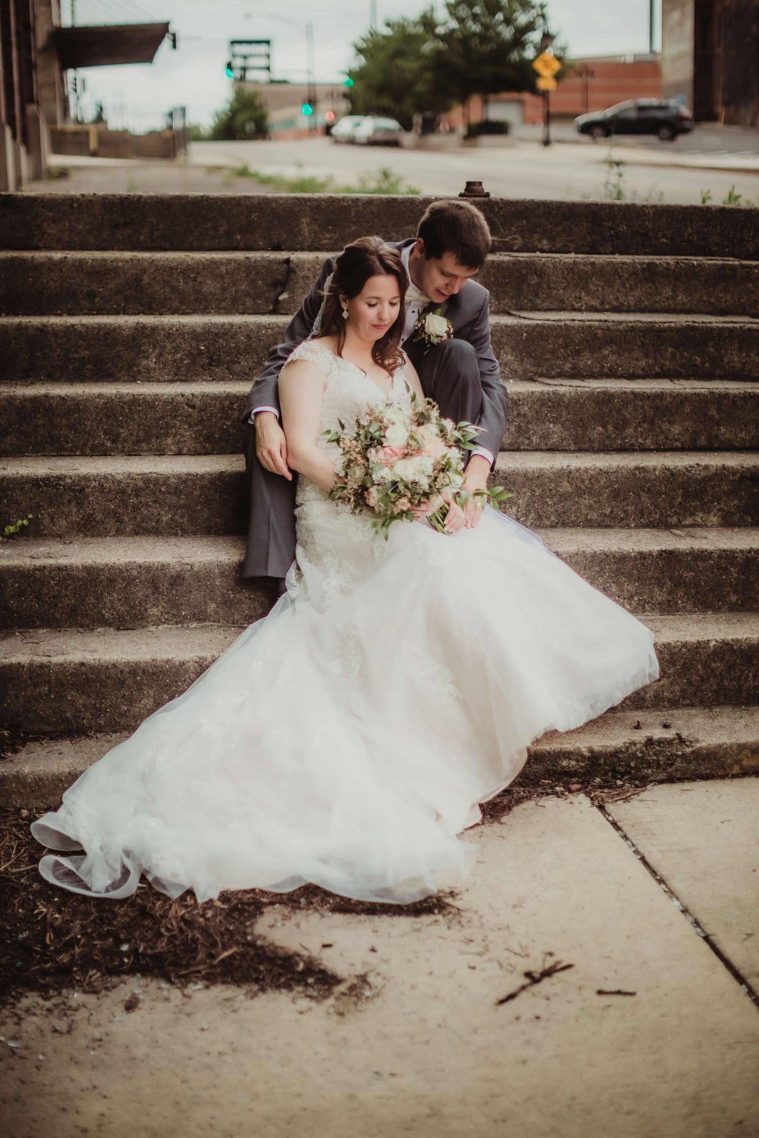A couple in wedding attire sitting on urban concrete steps with a city backdrop.