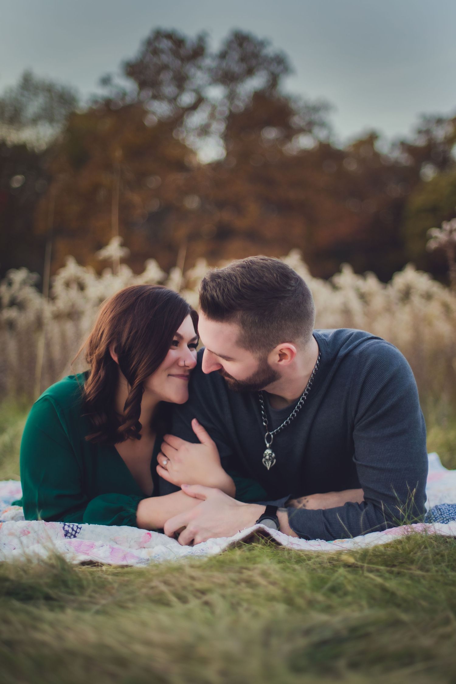 Couple lying on blanket sharing intimate moment in autumn field with tall grass and trees.