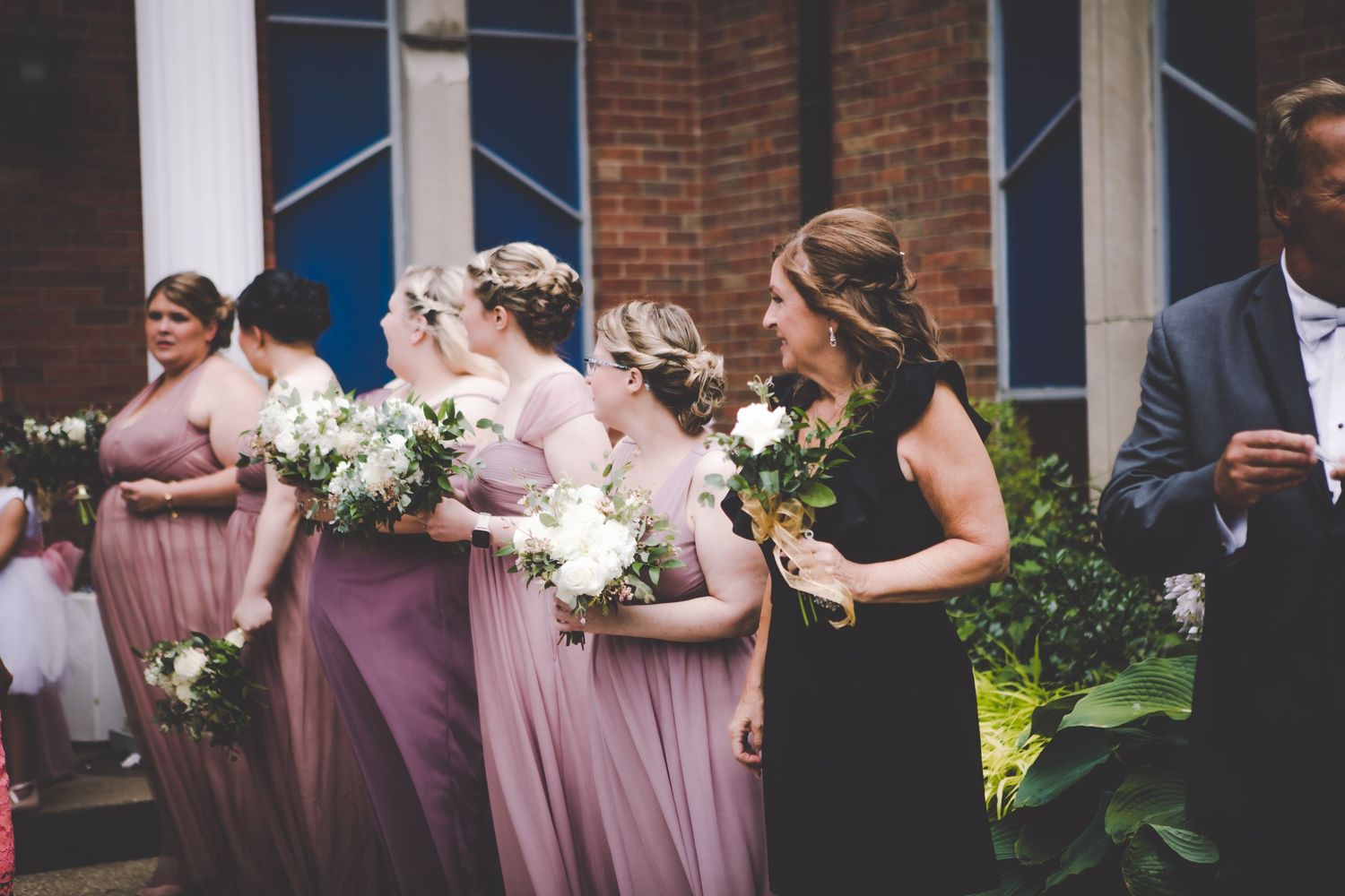 Bridesmaids in pink and mauve dresses hold white bouquets outside a brick building at a wedding ceremony.
