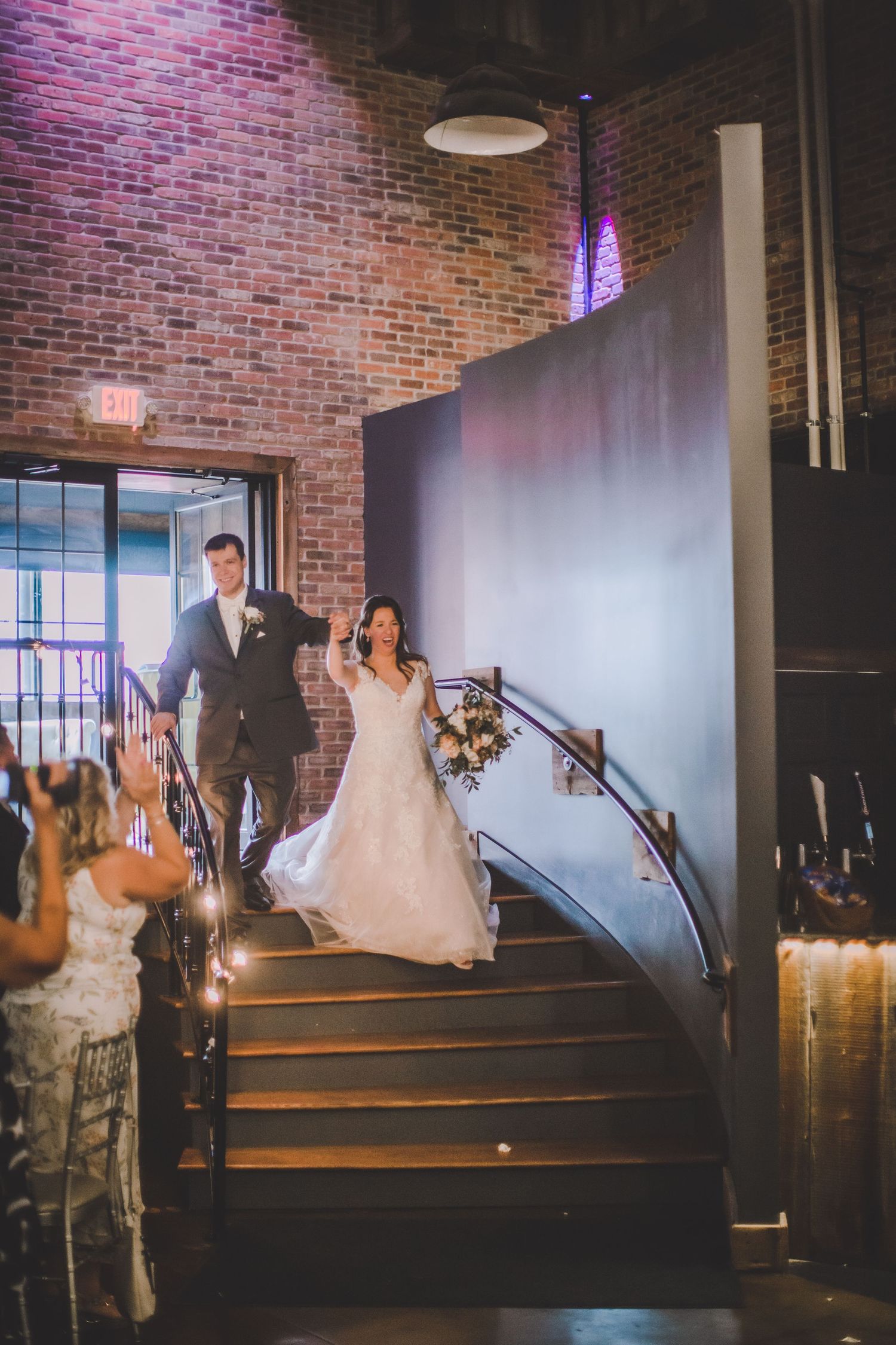 A newlywed couple makes their grand entrance down stairs at a rustic brick venue with dramatic lighting.