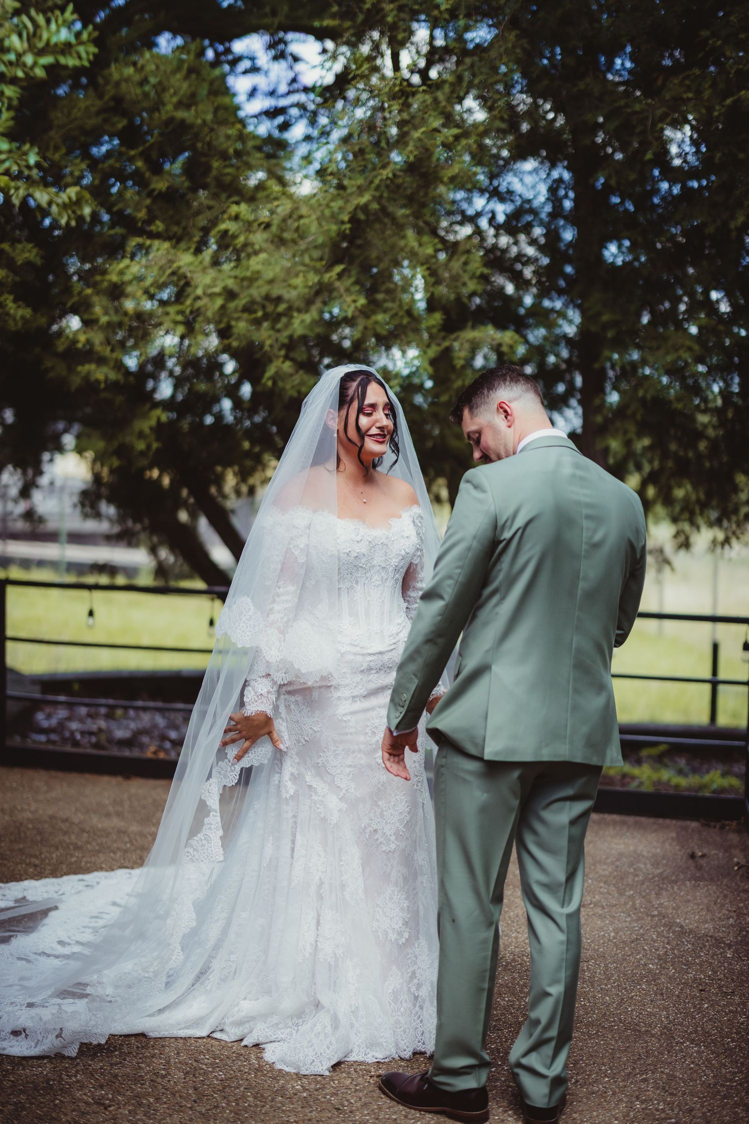 Bride in lace gown and veil holds hands with groom in sage suit outdoors near trees.