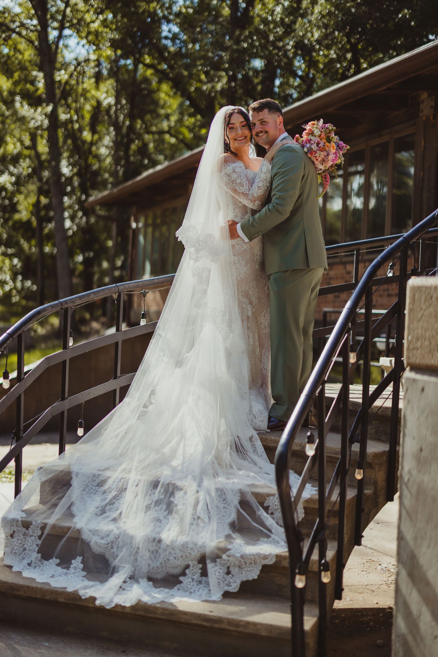 Bride in lace gown with veil and groom in sage suit embrace on stone steps surrounded by trees.