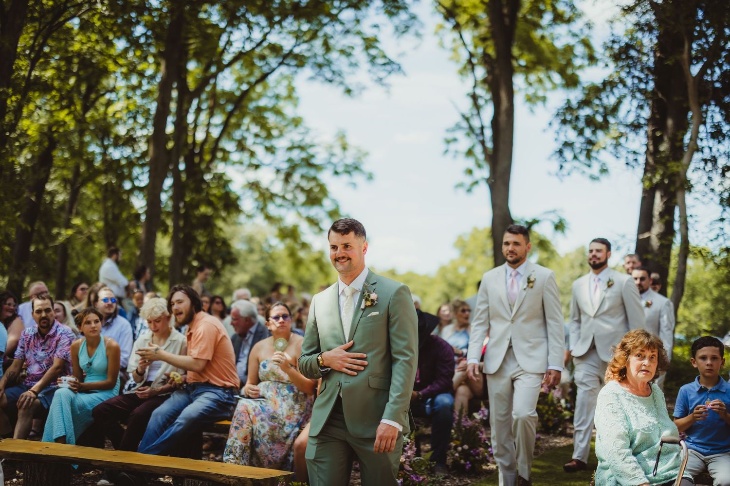 Groom in sage green suit walks down outdoor wedding aisle surrounded by seated guests and lush trees.