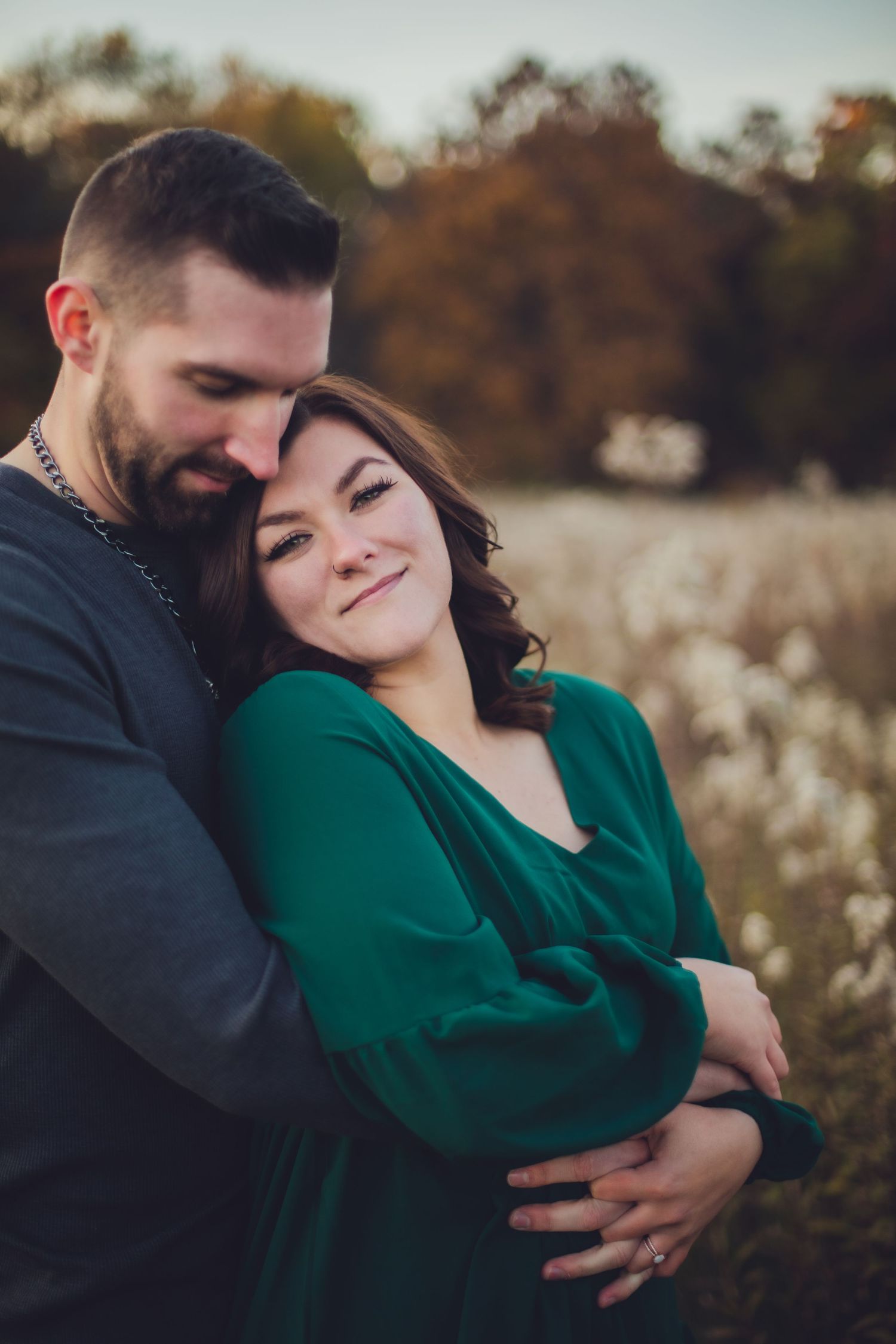 A romantic couple embraces in a field during autumn, with her wearing an emerald green dress and him in dark grey.