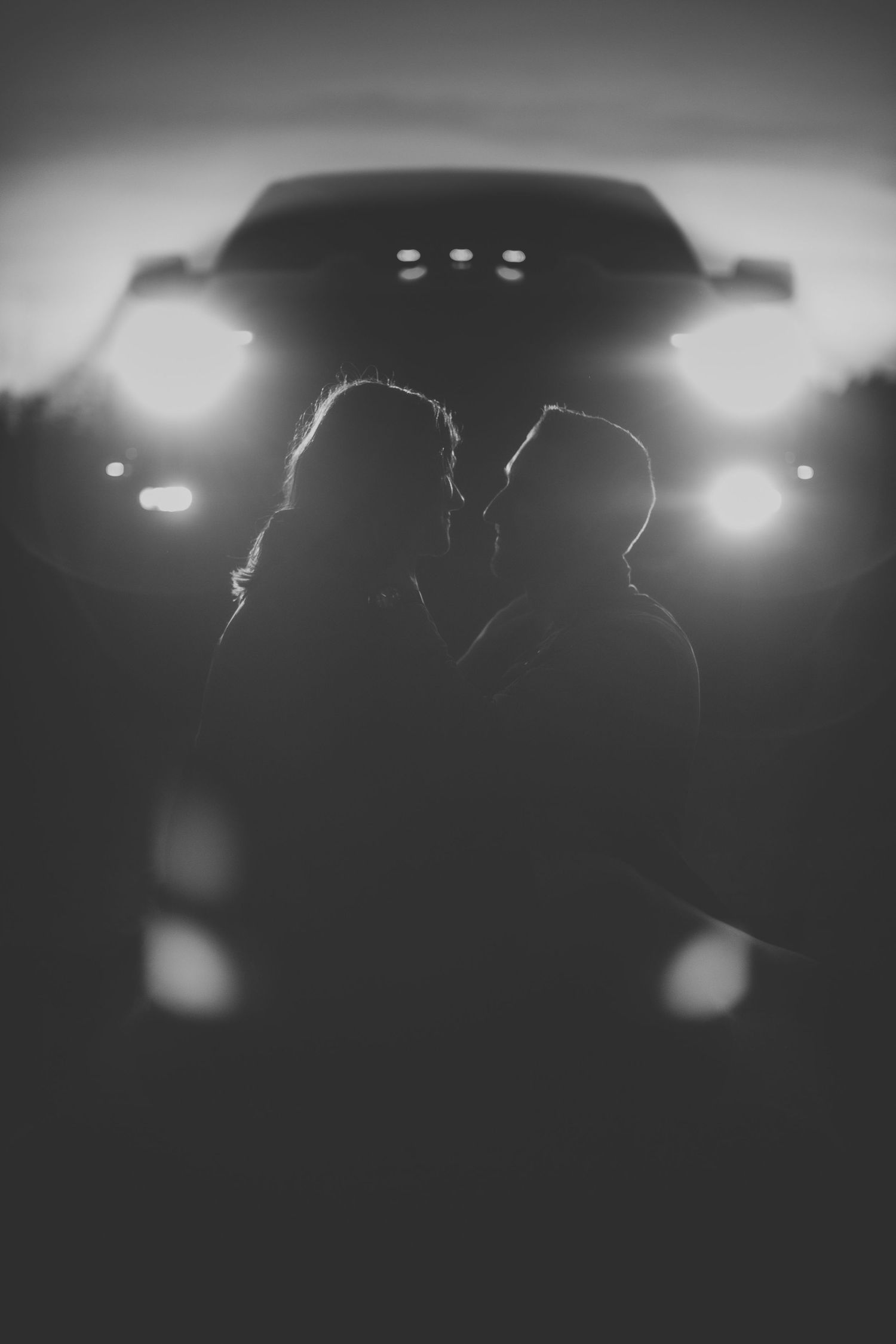 Silhouetted couple against car headlights in moody black and white composition.