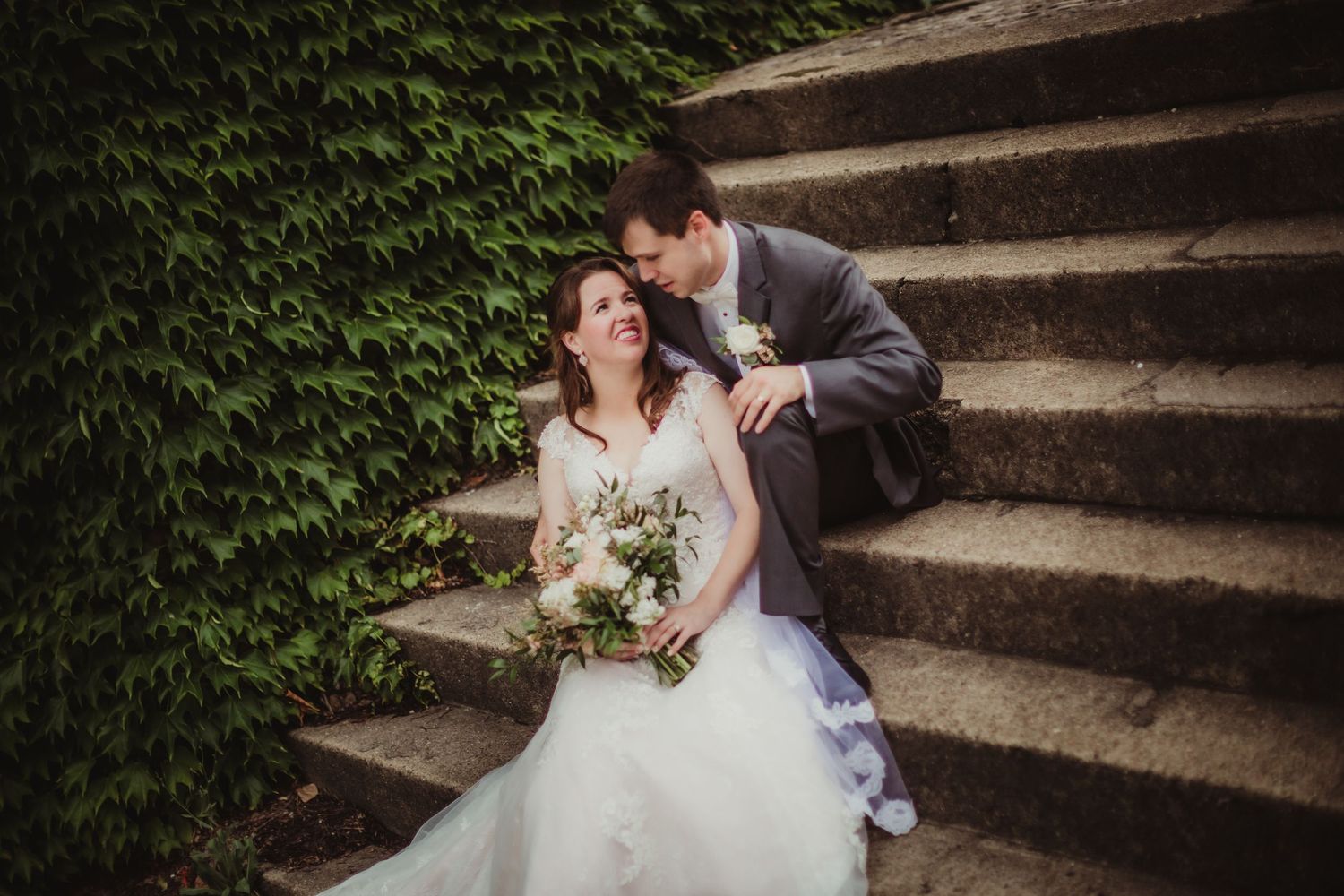 Newlyweds share an intimate moment on garden steps with a floral backdrop.