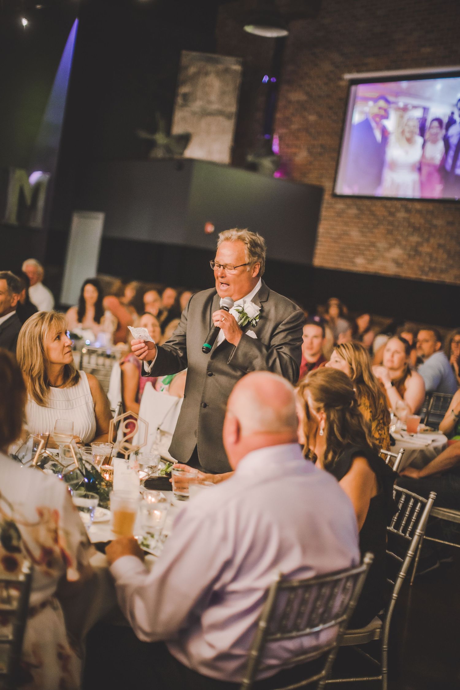 A speaker delivers a toast during a wedding reception while guests listen attentively at decorated tables.