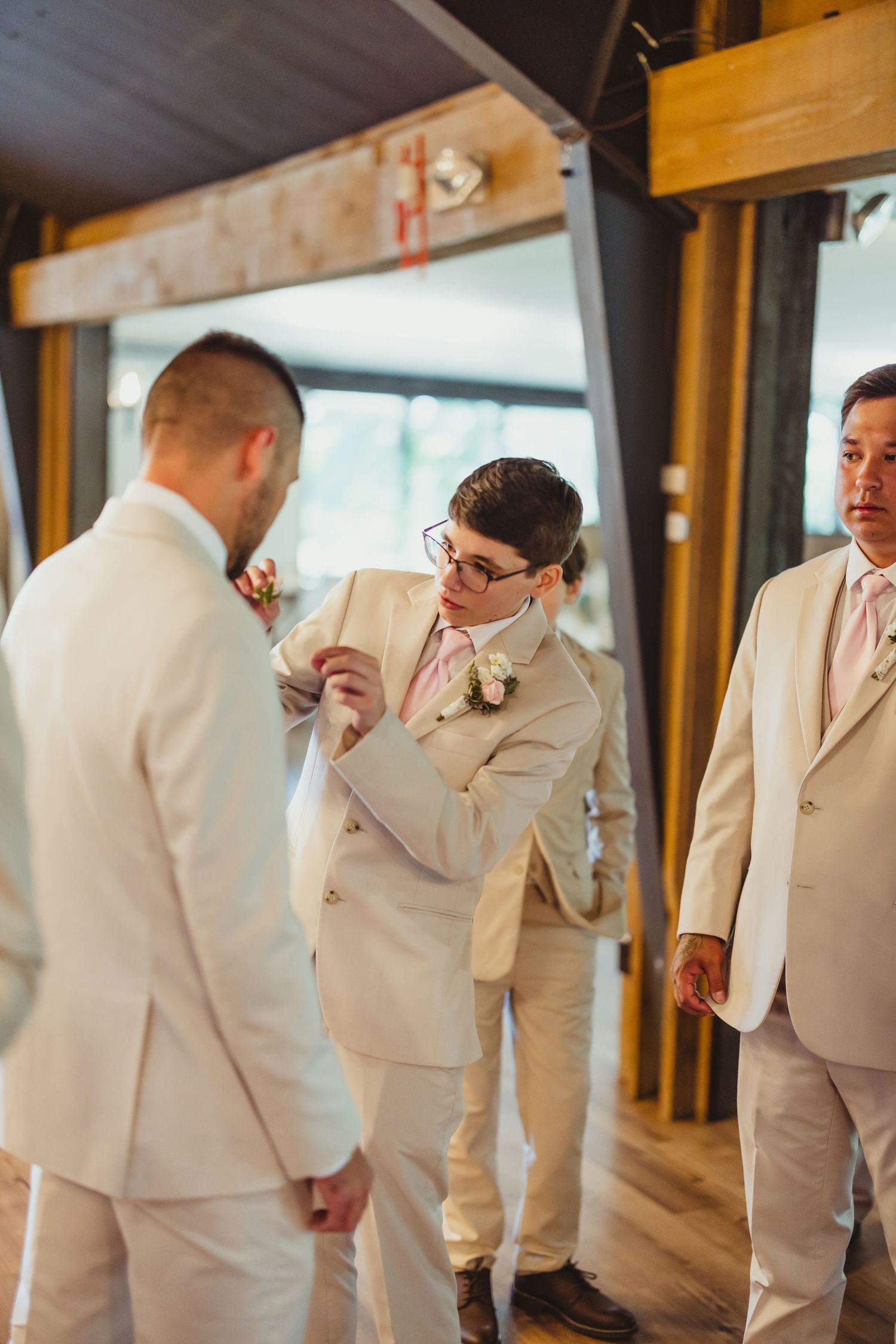 Groomsmen in beige suits with pink ties help each other with boutonnieres inside a rustic wedding venue.