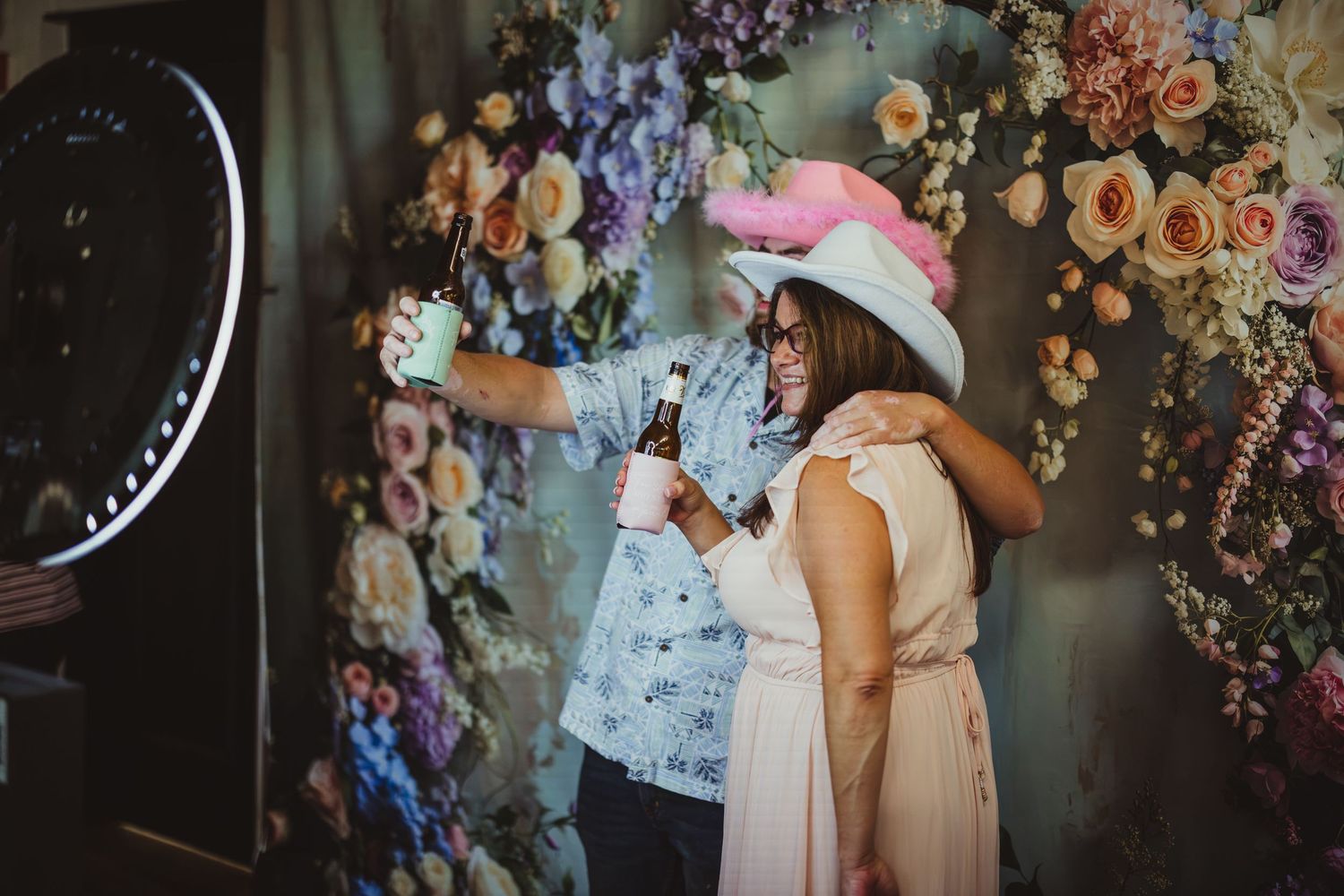 Two guests pose for a selfie in front of a floral wall backdrop at an event, one wearing a white hat with pink feathers.