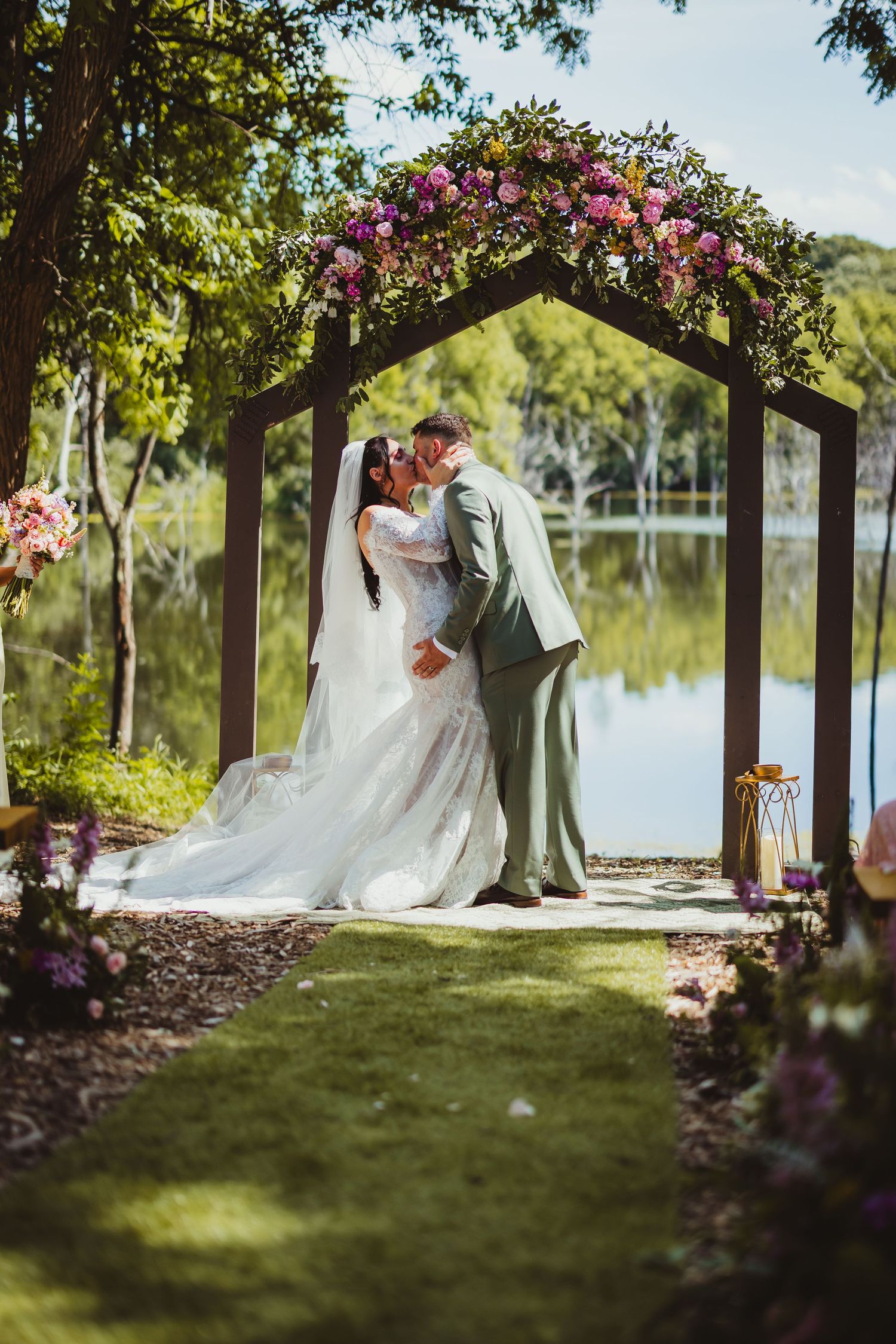 Bride and groom share first kiss under floral arch by lakeside at romantic outdoor wedding ceremony.