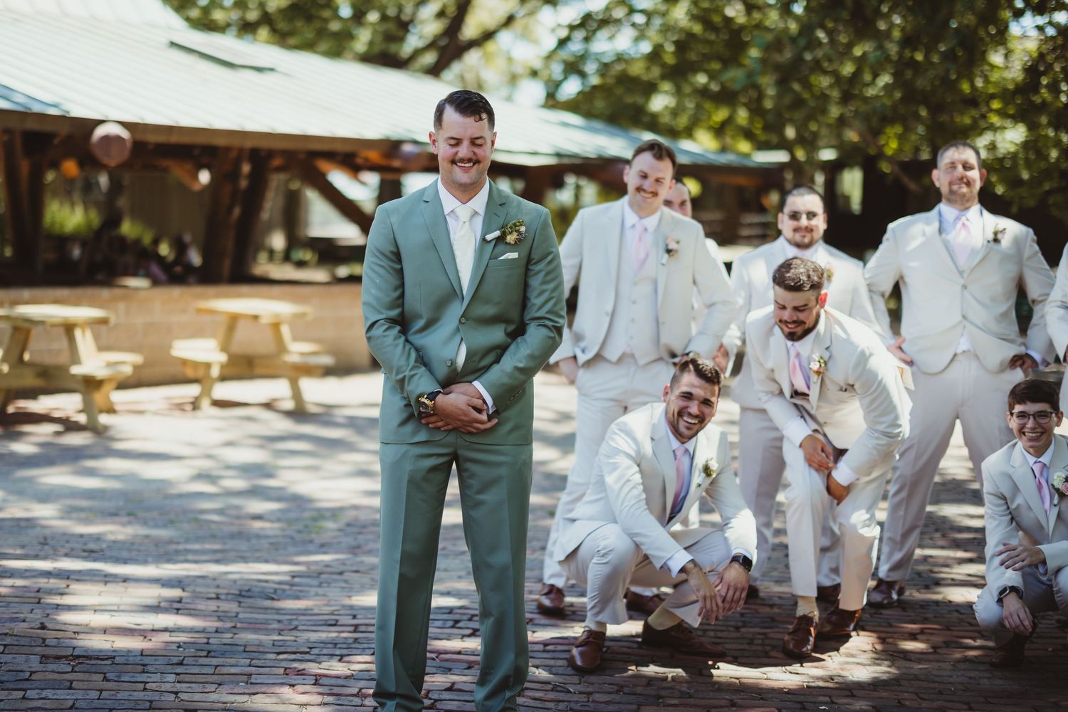 Groom in sage green suit laughing with groomsmen in light gray suits outdoors at a wedding venue.