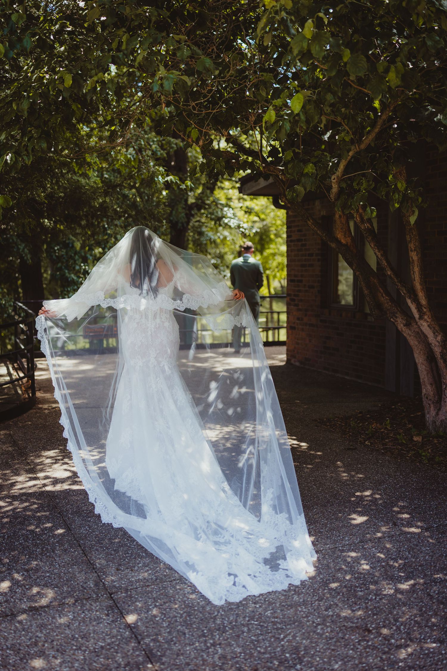 Bride in lace wedding gown with flowing veil in sunlit garden, groom standing in background among green trees.