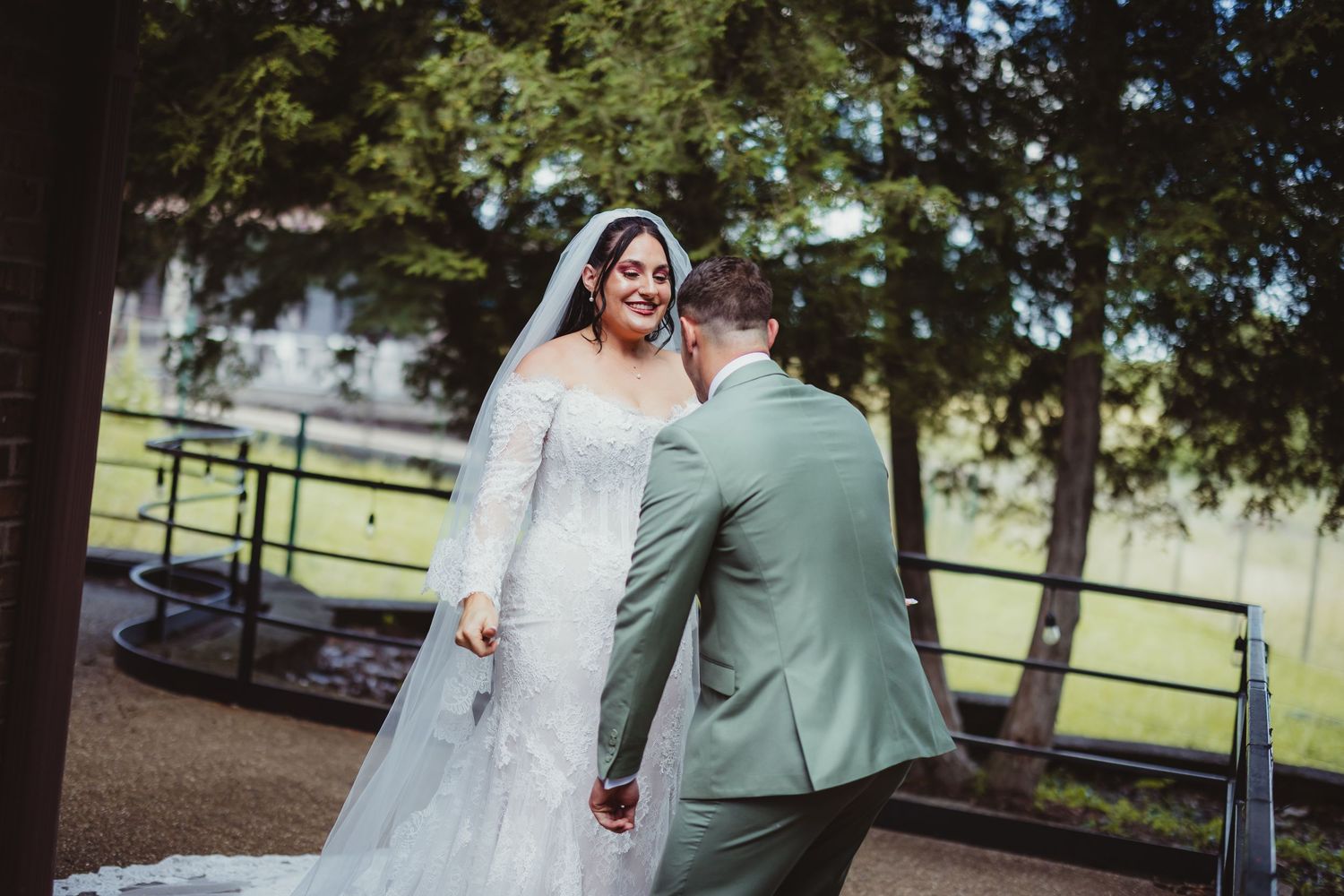 Bride in white gown and veil smiles at groom in sage green suit outdoors near trees.