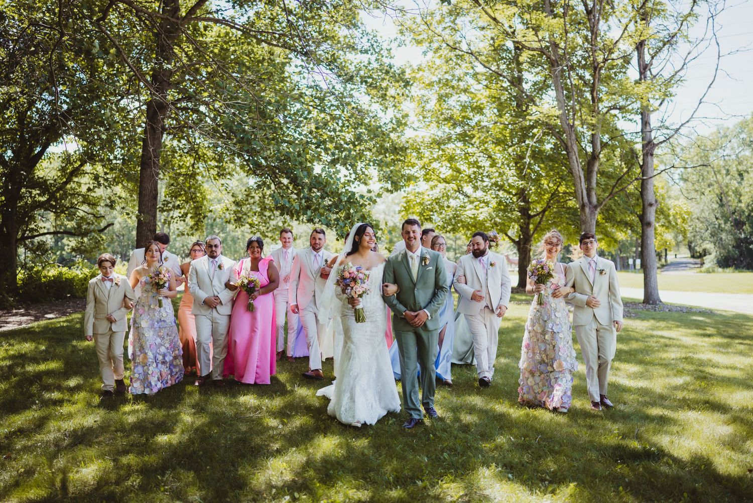 Wedding party walks outdoors on lush green lawn, bride in white gown, bridesmaids in pink and floral dresses, groomsmen in tan suits.