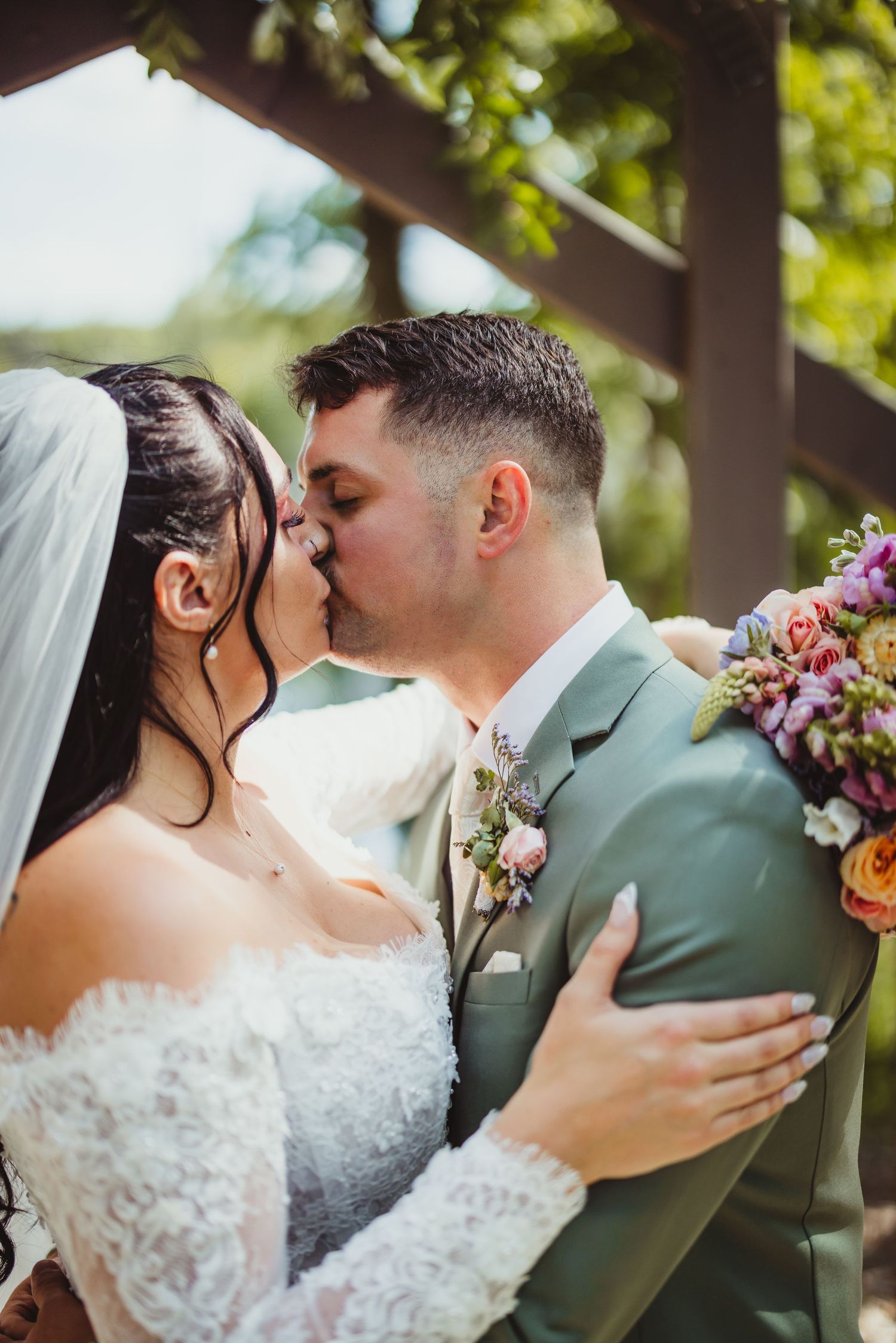 Bride and groom sharing first kiss at outdoor wedding ceremony, bride holding vibrant floral bouquet.