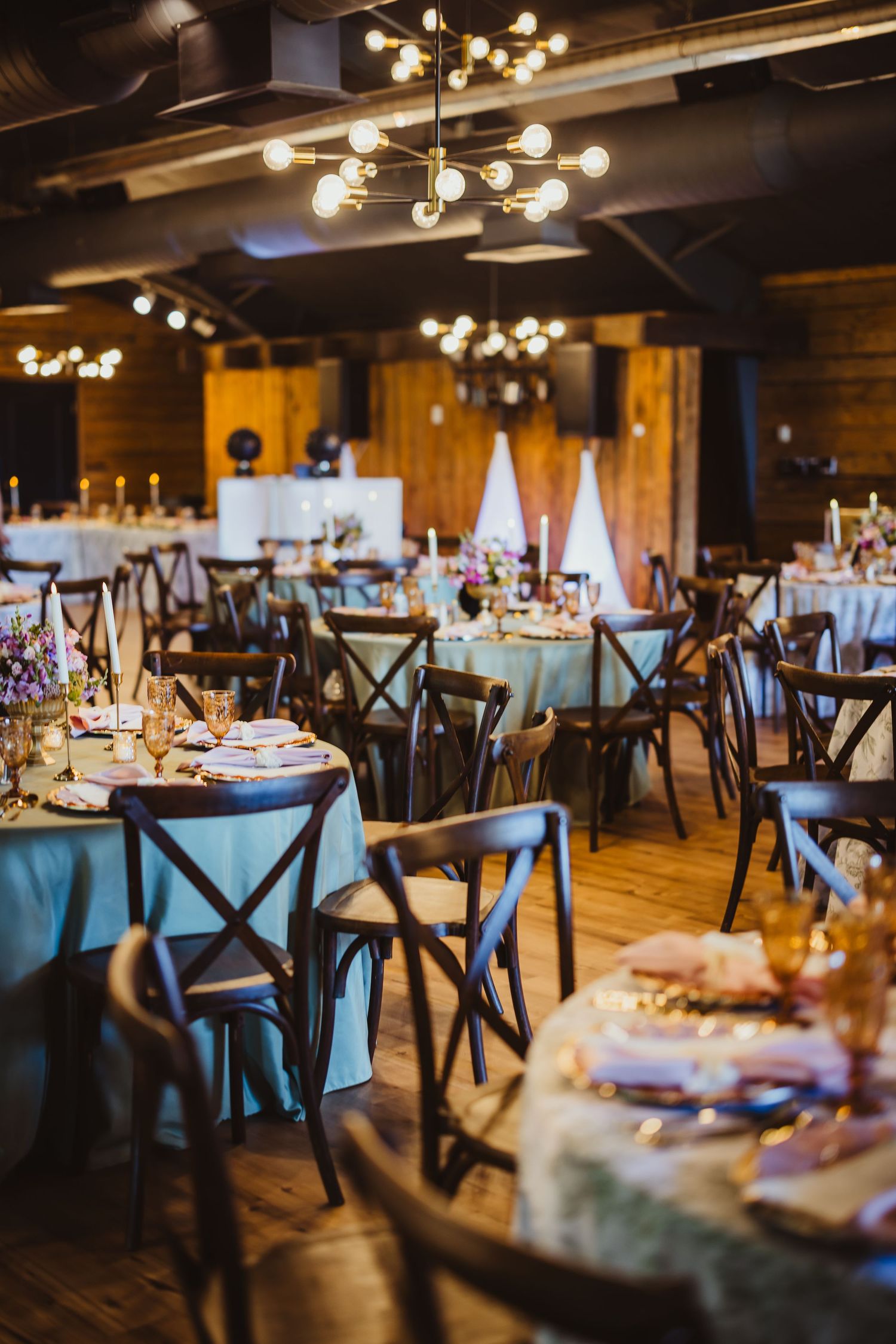 Elegant rustic wedding reception hall with blue tablecloths, dark cross-back chairs, and warm Edison bulb chandeliers.