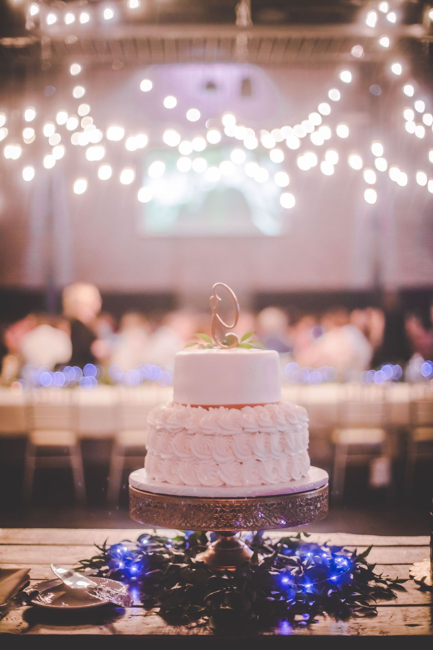 Two-tiered white wedding cake with string lights and blue accent lighting in reception hall.