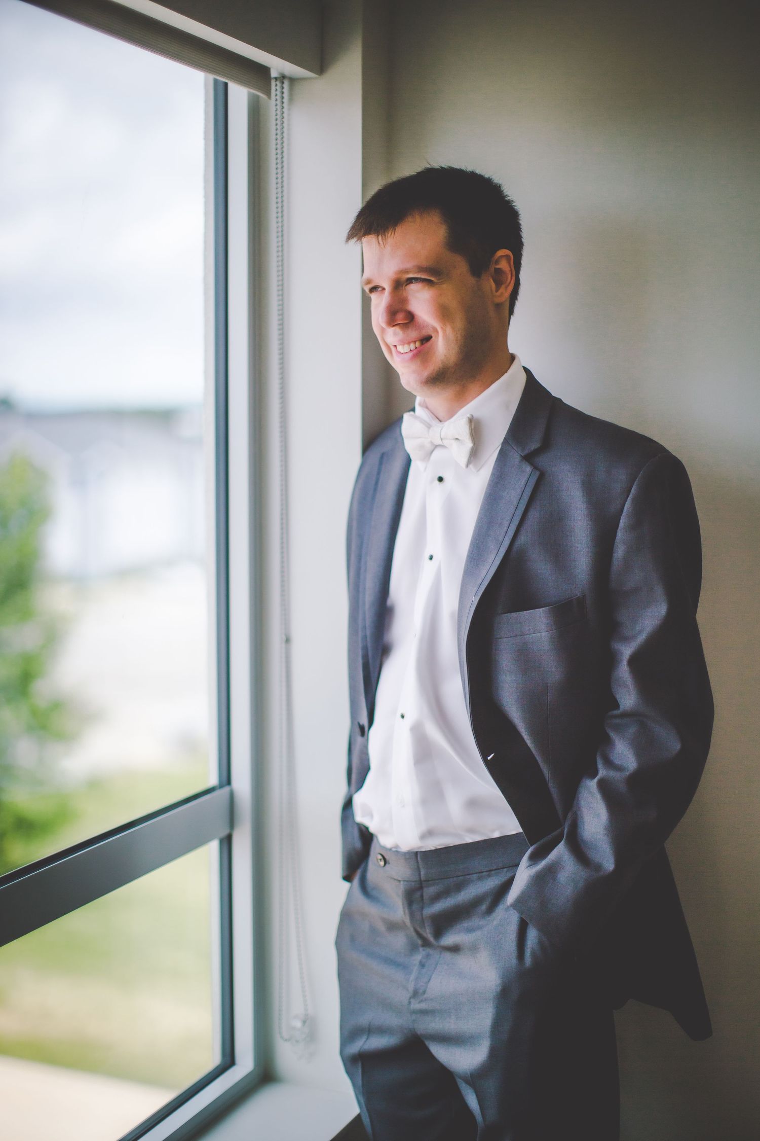 Groom in formal grey suit and white bowtie smiles while looking through window in natural lighting.