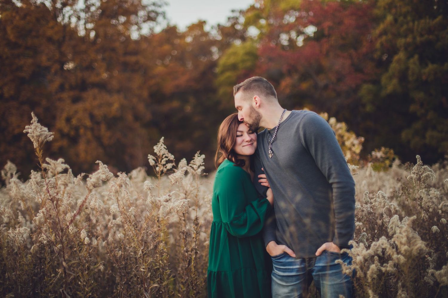 Autumn engagement photo of couple embracing in field of tall grass with fall foliage.
