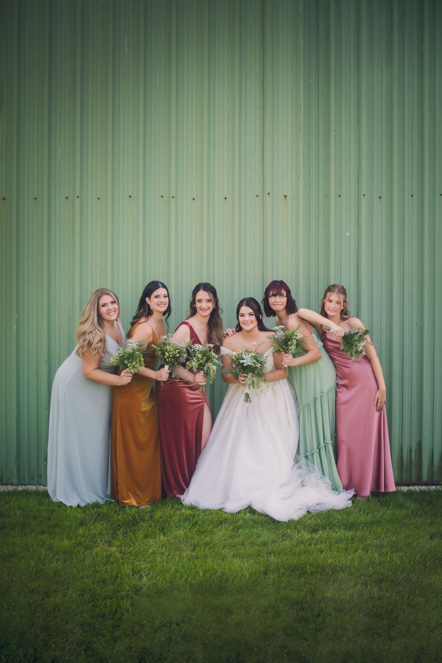 Wedding party in pastel dresses arranged artistically against green metal wall backdrop.