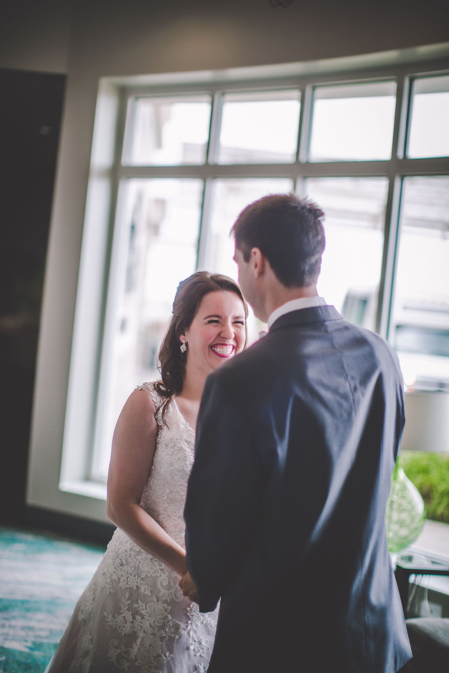 A couple shares laughter during an intimate wedding moment by large windows.