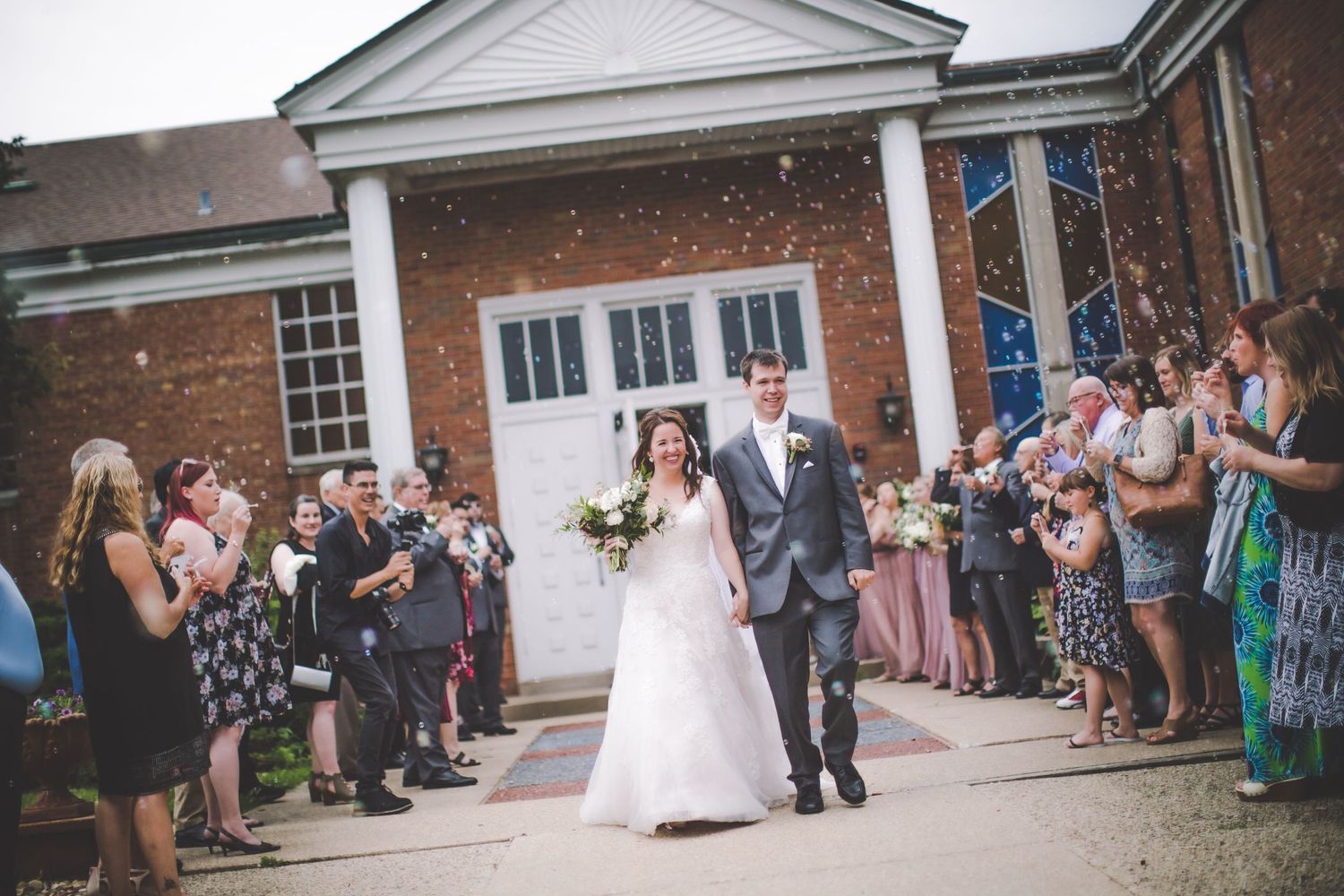 Wedding party and guests gathered outside colonial-style brick building entrance.