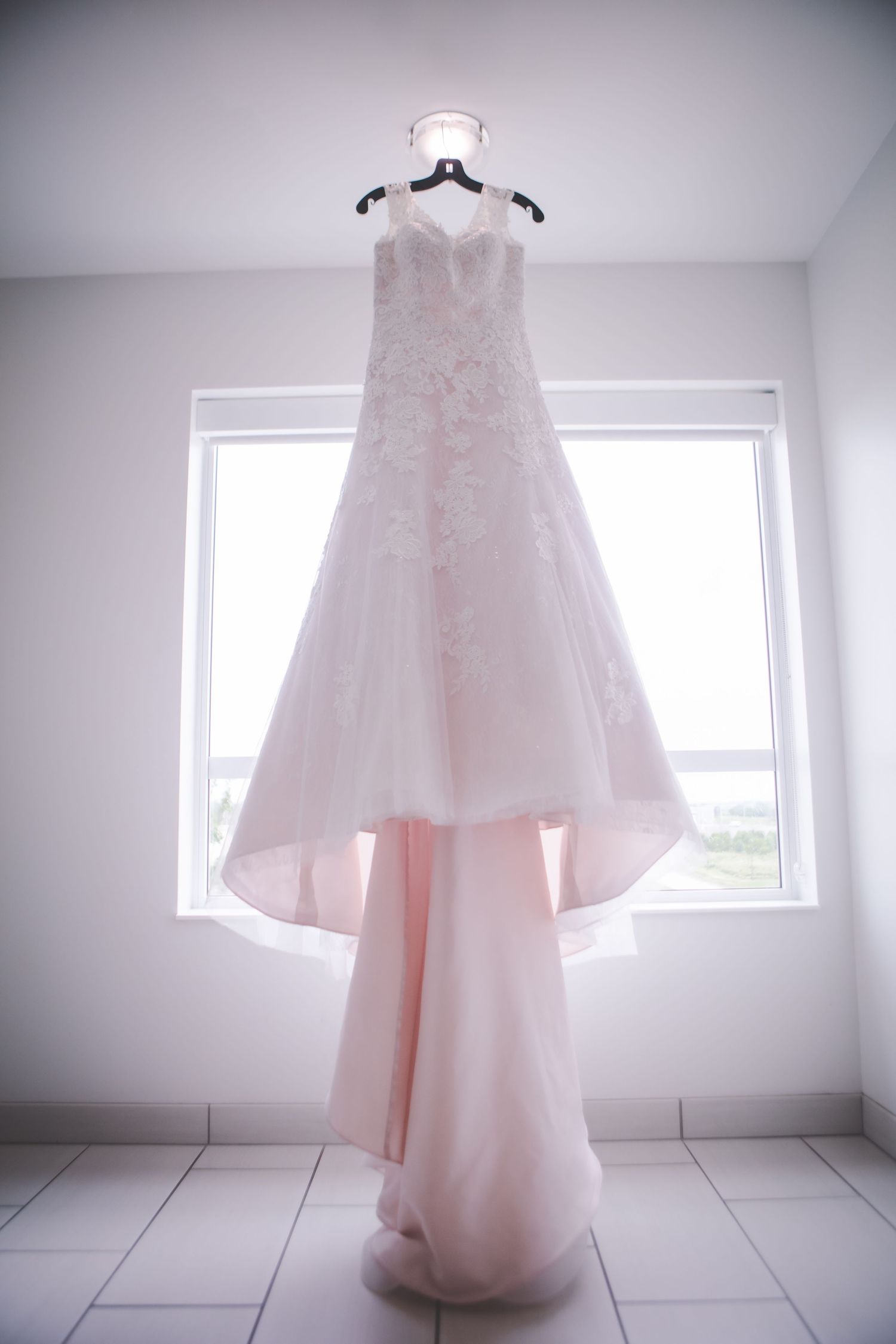 Ethereal blush wedding dress hanging in white room with natural window light.