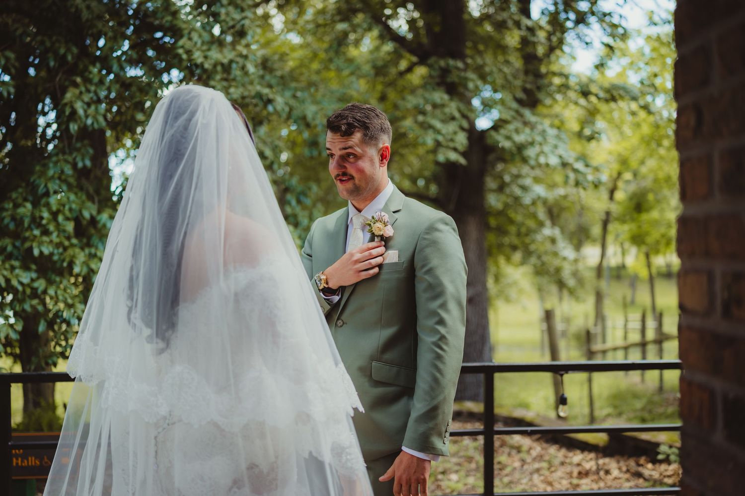 Emotional groom in sage green suit reacts to first look with bride in white veil at outdoor wedding.