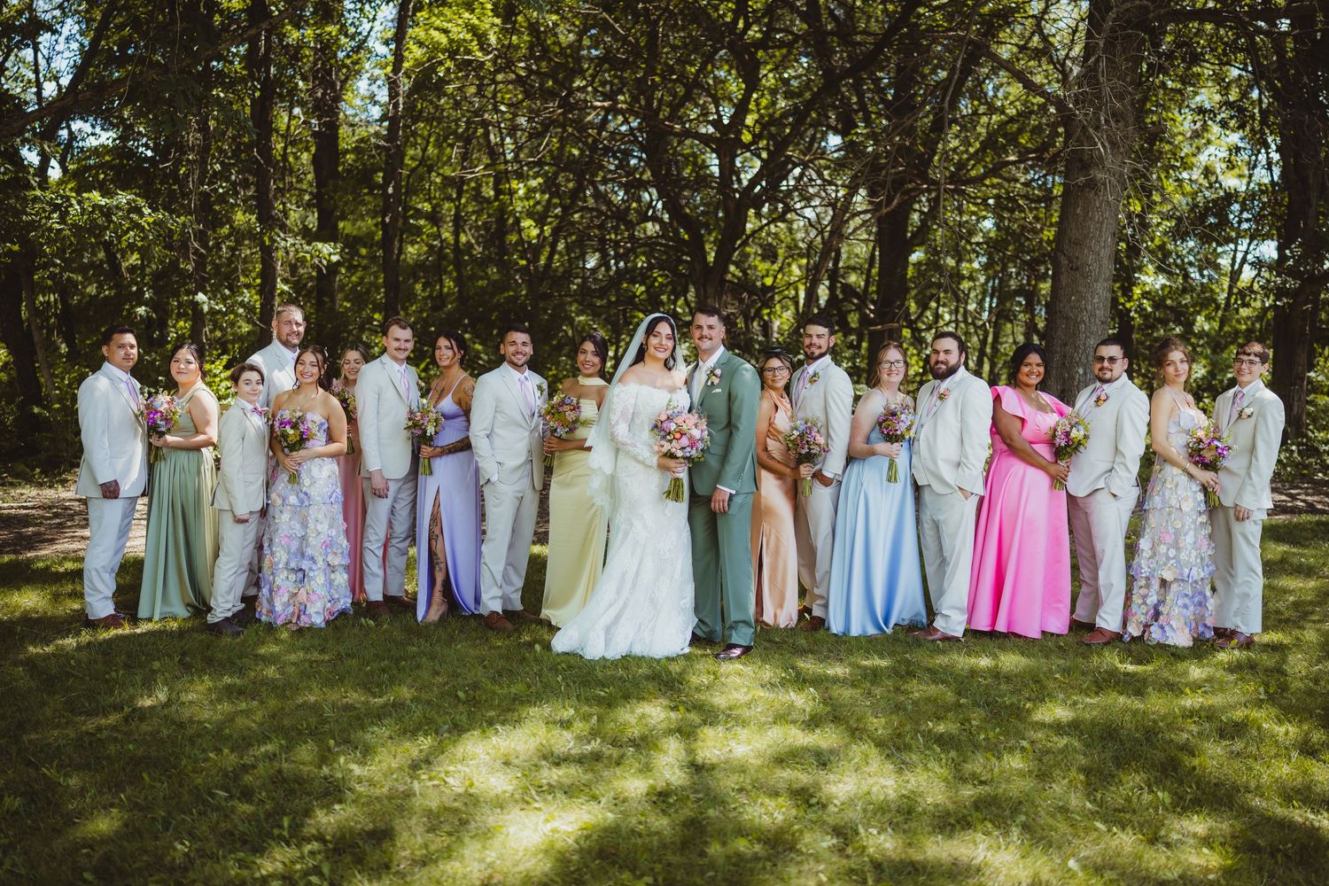 Wedding party poses outdoors in colorful gowns and white suits, surrounded by lush green trees on a sunny day.