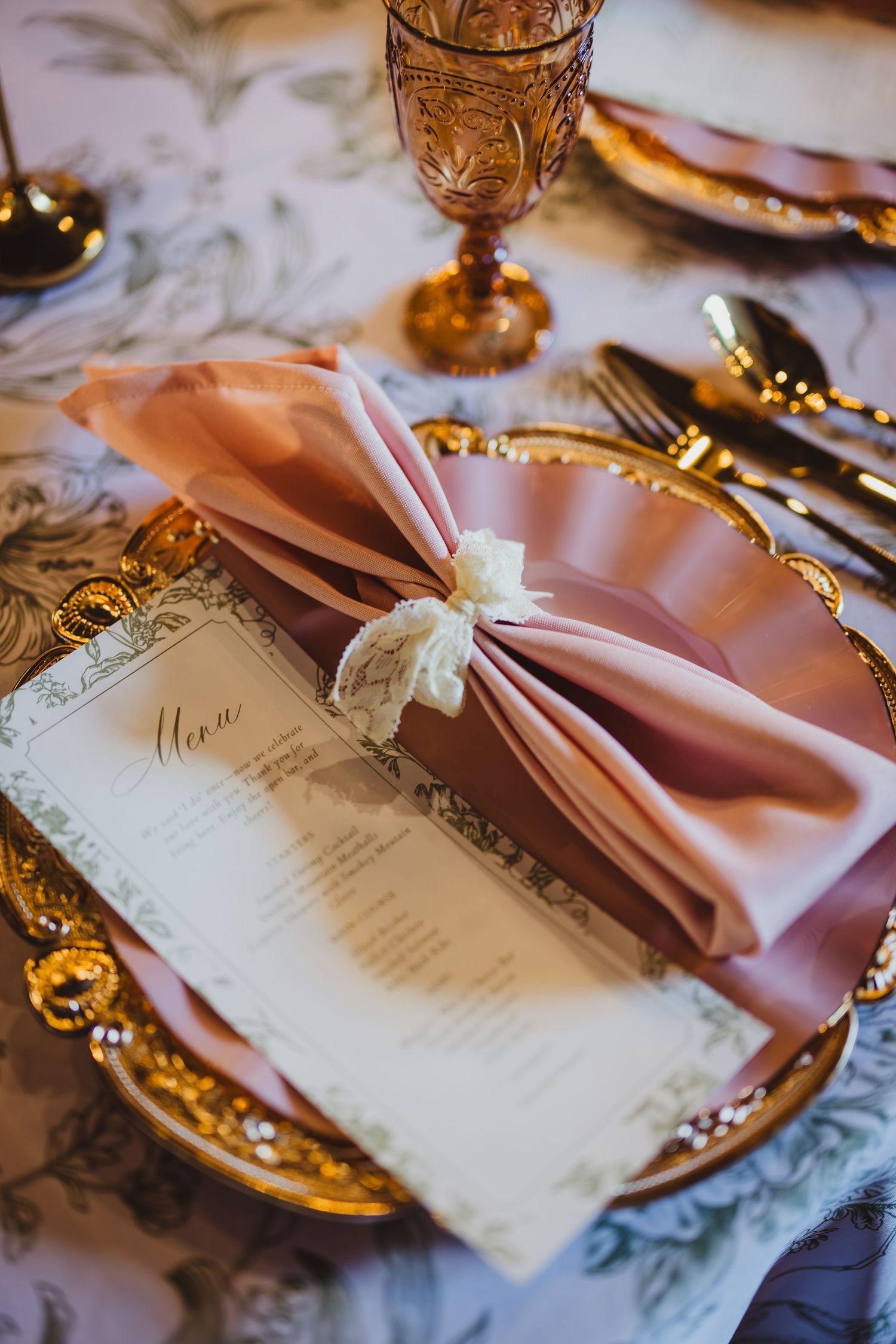 Elegant wedding table setting with blush pink napkin, gold charger plate, menu card, and amber goblet on floral tablecloth.