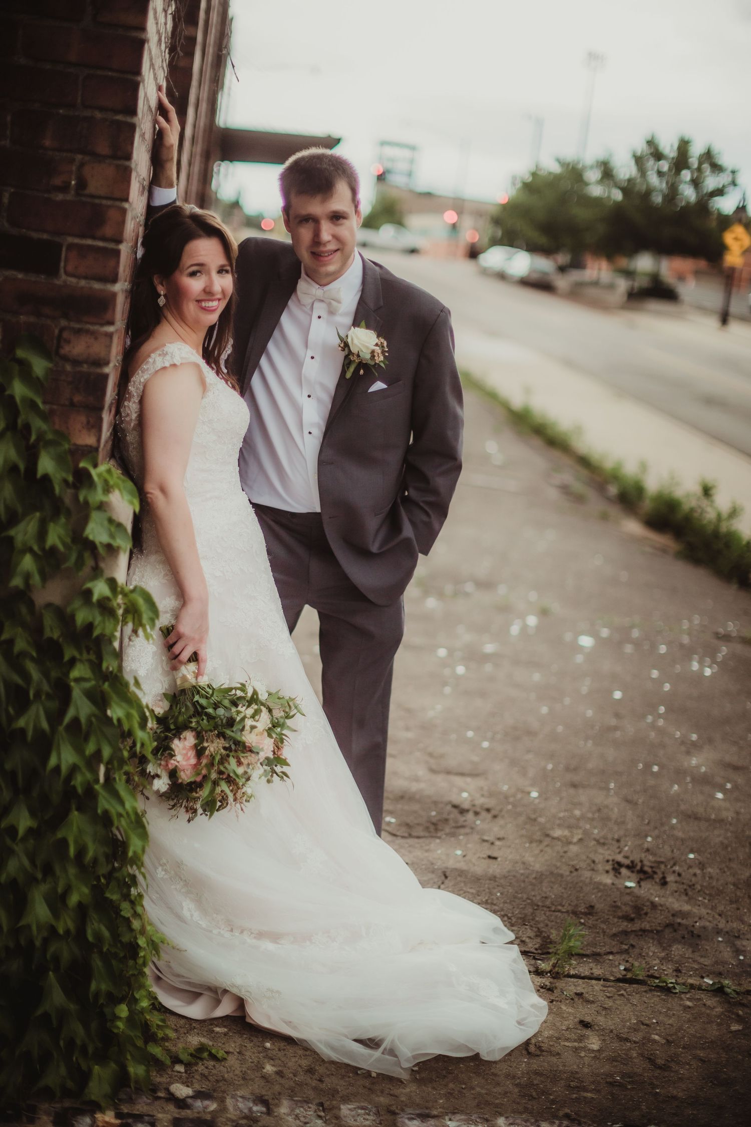 Wedding couple poses by vintage brick wall with trailing vines, bride holding natural floral bouquet.