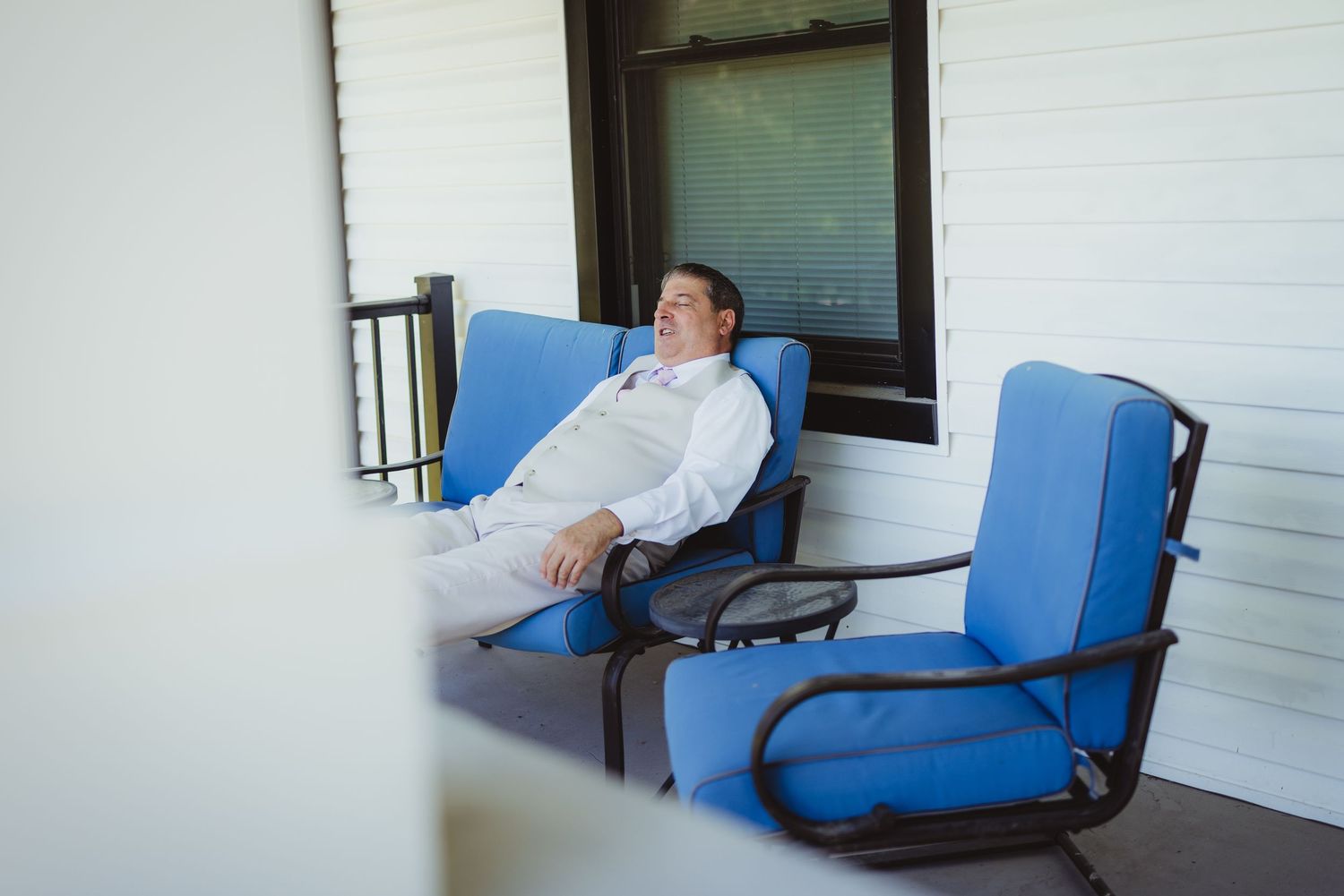 Man relaxing in blue patio chair on covered porch, wearing white shirt, eyes closed, resting peacefully outdoors.