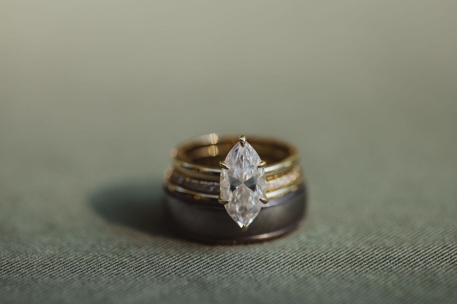 Marquise diamond engagement ring with gold wedding band and dark wood ring on sage green fabric background.