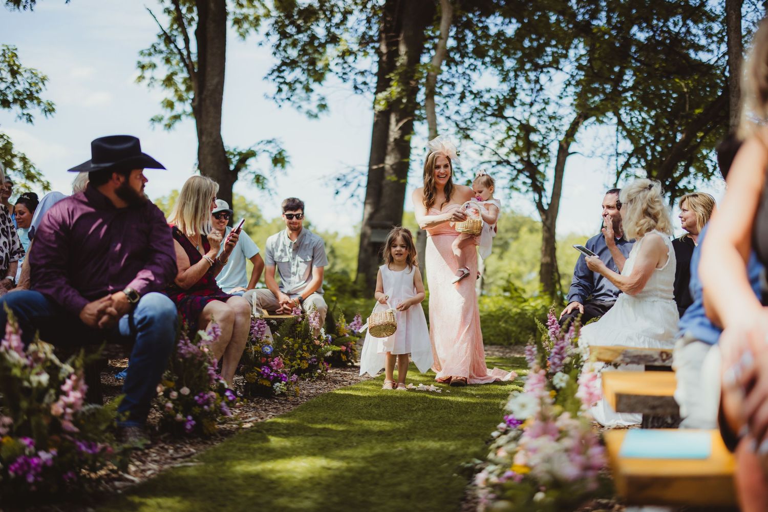 Outdoor wedding ceremony with guests seated along a flower-lined aisle as a bridesmaid carries a baby toward the altar.