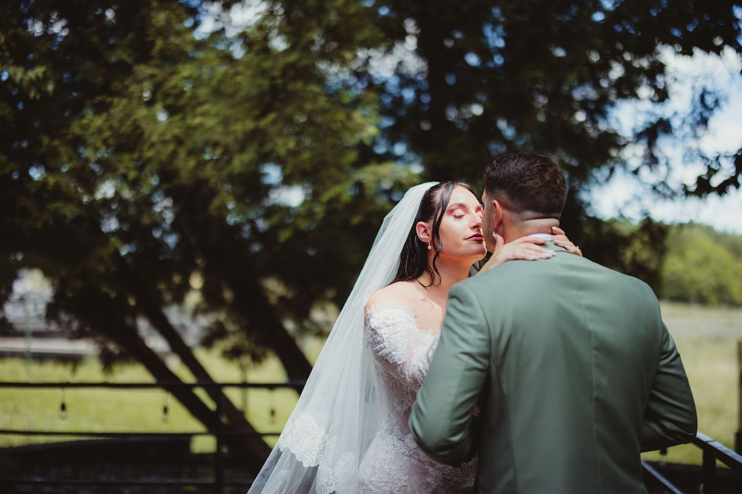 A bride in a white gown and veil shares a romantic kiss with a groom in a sage suit outdoors.