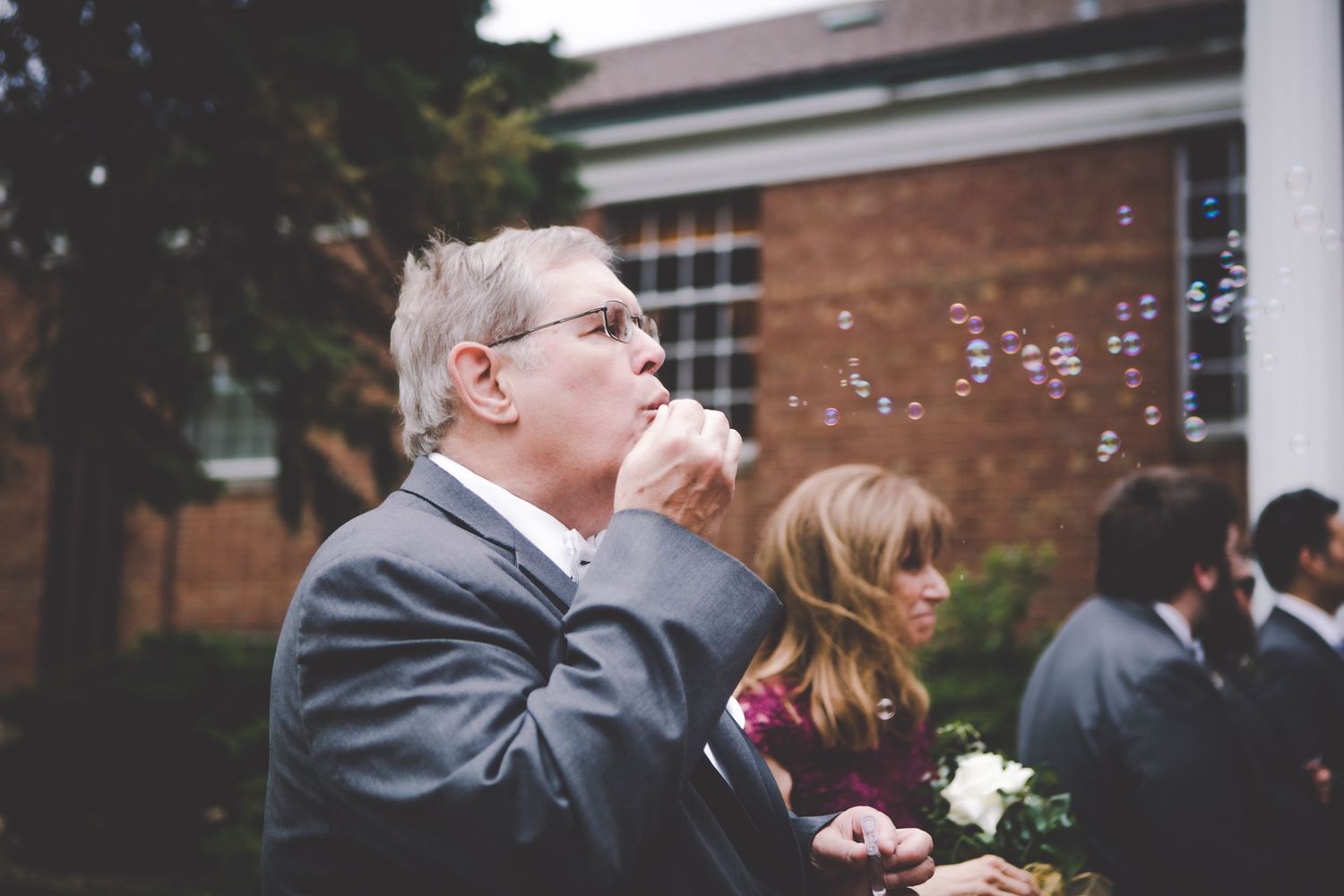 An older gentleman in a gray suit blows bubbles during an outdoor wedding celebration.