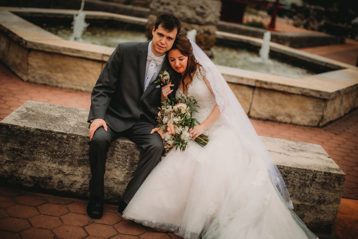 Wedding couple sits together on brick steps with ornate fountain in background.