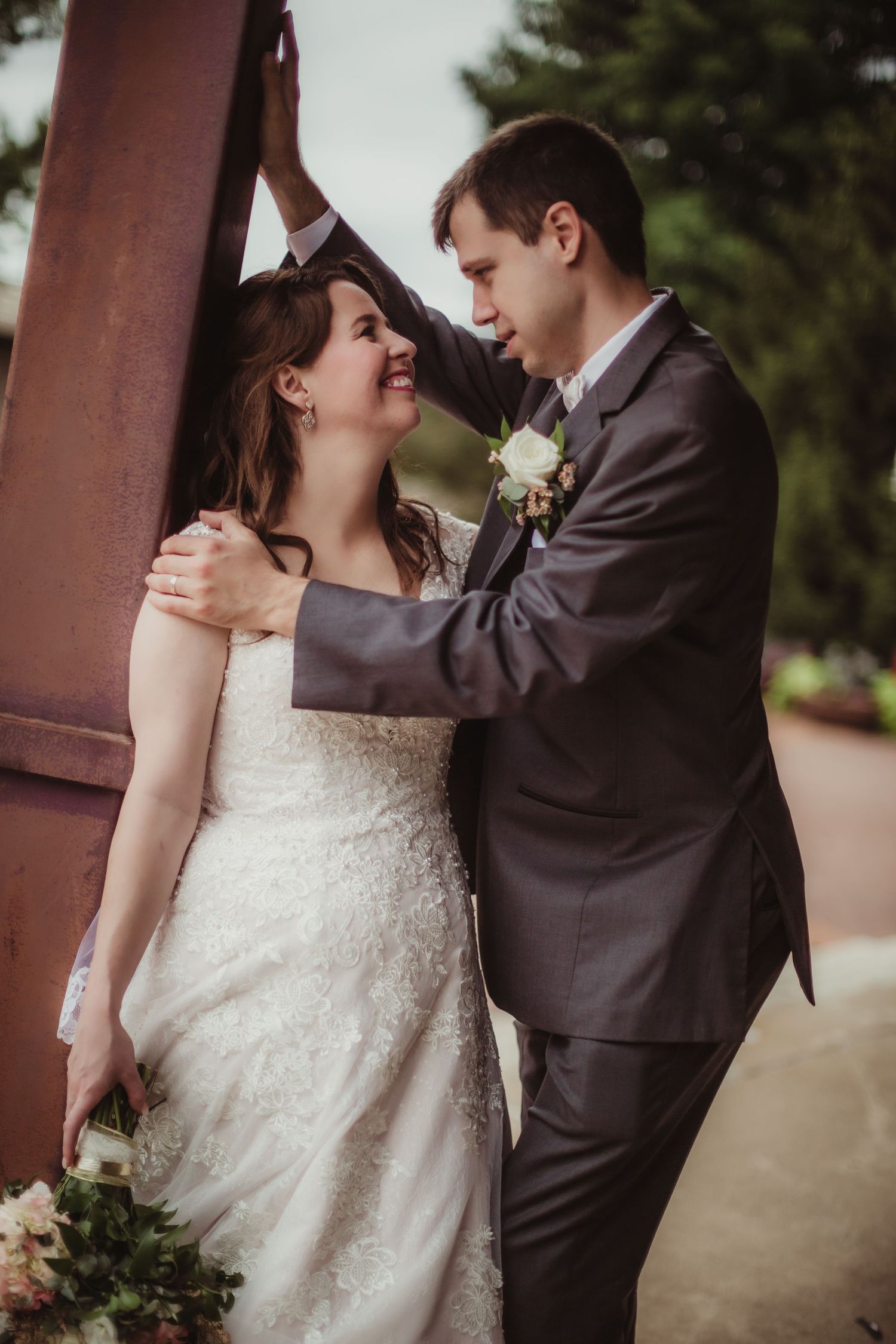 A romantic wedding couple embraces near a rustic metal pillar with the bride in white lace and groom in charcoal suit.
