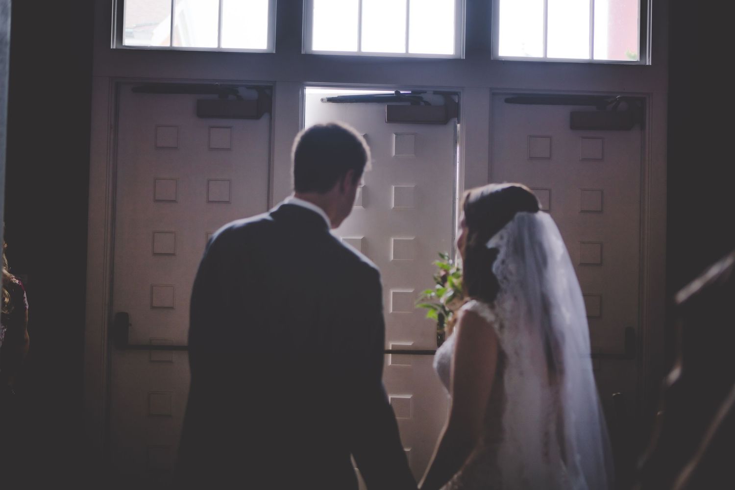 A couple stands in silhouette against bright church windows during their wedding ceremony.