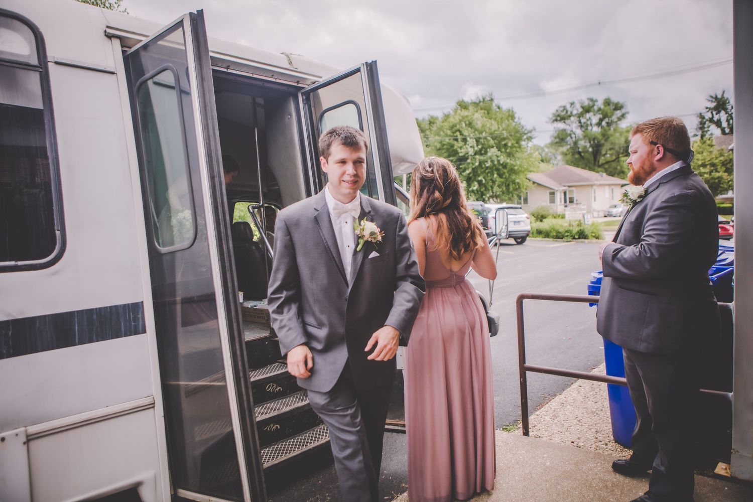 Wedding party gathering around white shuttle bus parked outside venue.
