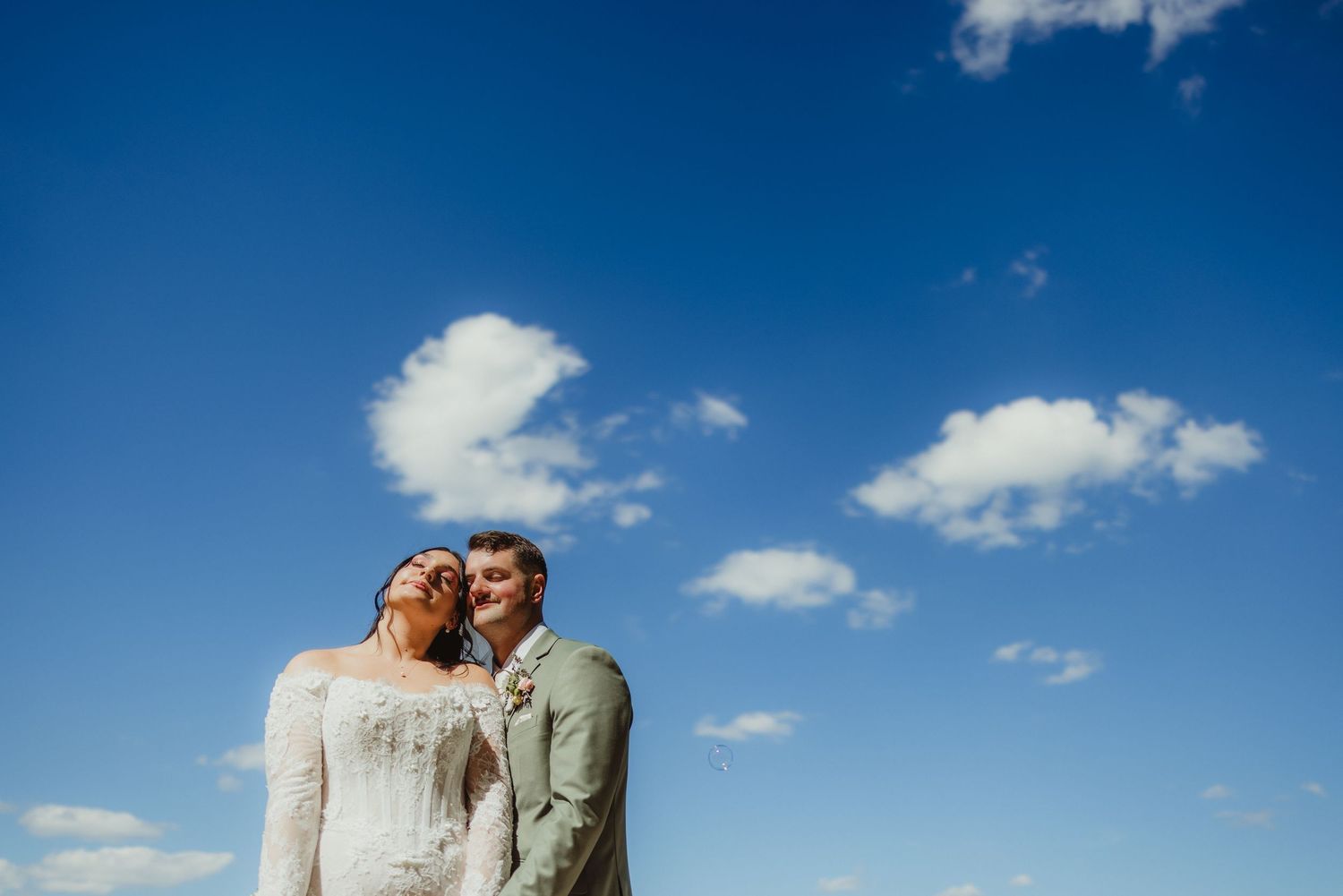 A joyful wedding couple embraces under a bright blue sky with scattered white clouds on their special day.