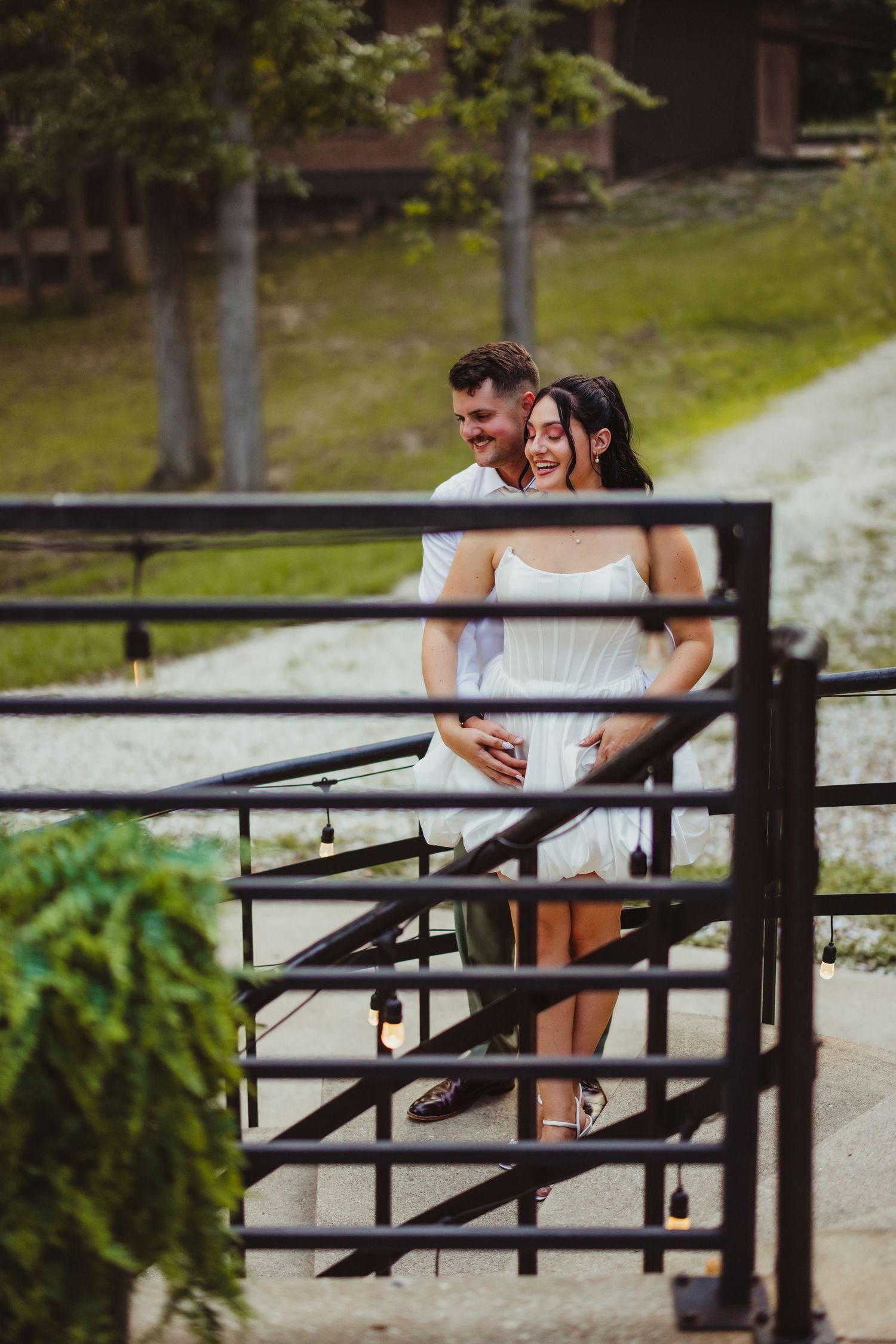 Happy couple embracing and laughing together on a rustic outdoor staircase with string lights at a countryside venue.