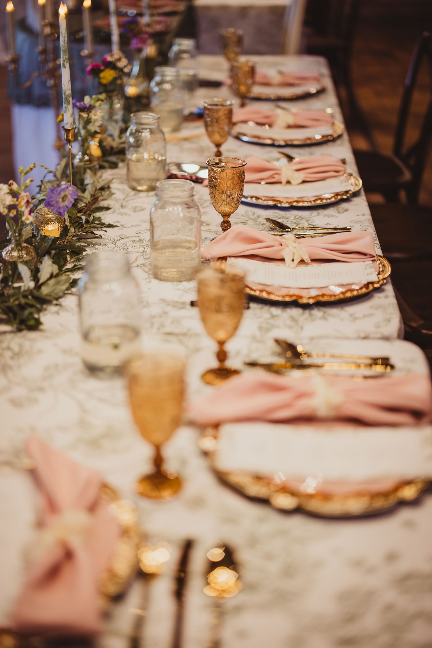 Detailed wedding banquet table with pink napkins, gold-rimmed plates, amber goblets, and floral greenery runner.