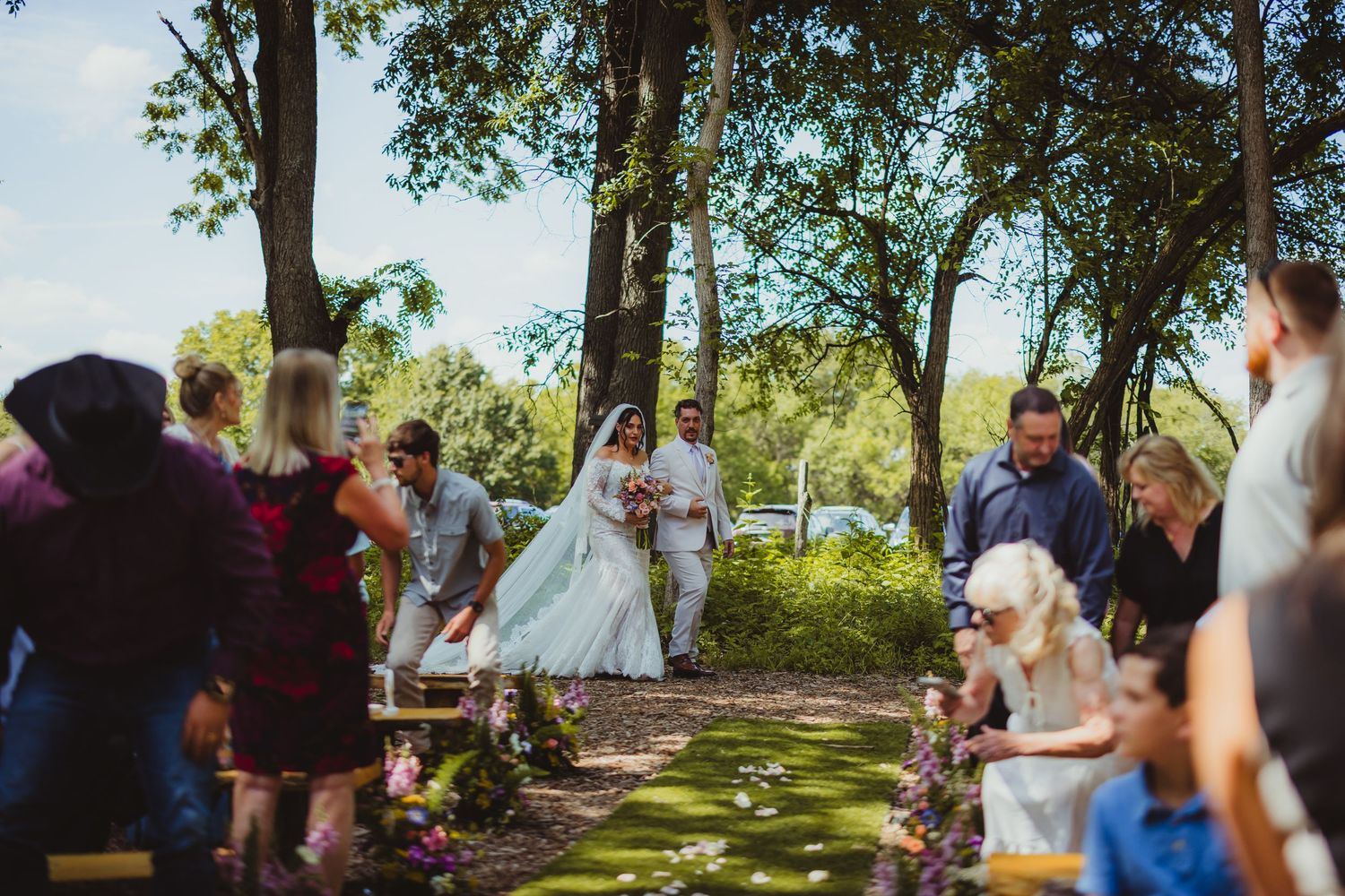 Bride and groom walk down outdoor aisle surrounded by guests and trees at a sunlit garden wedding ceremony.