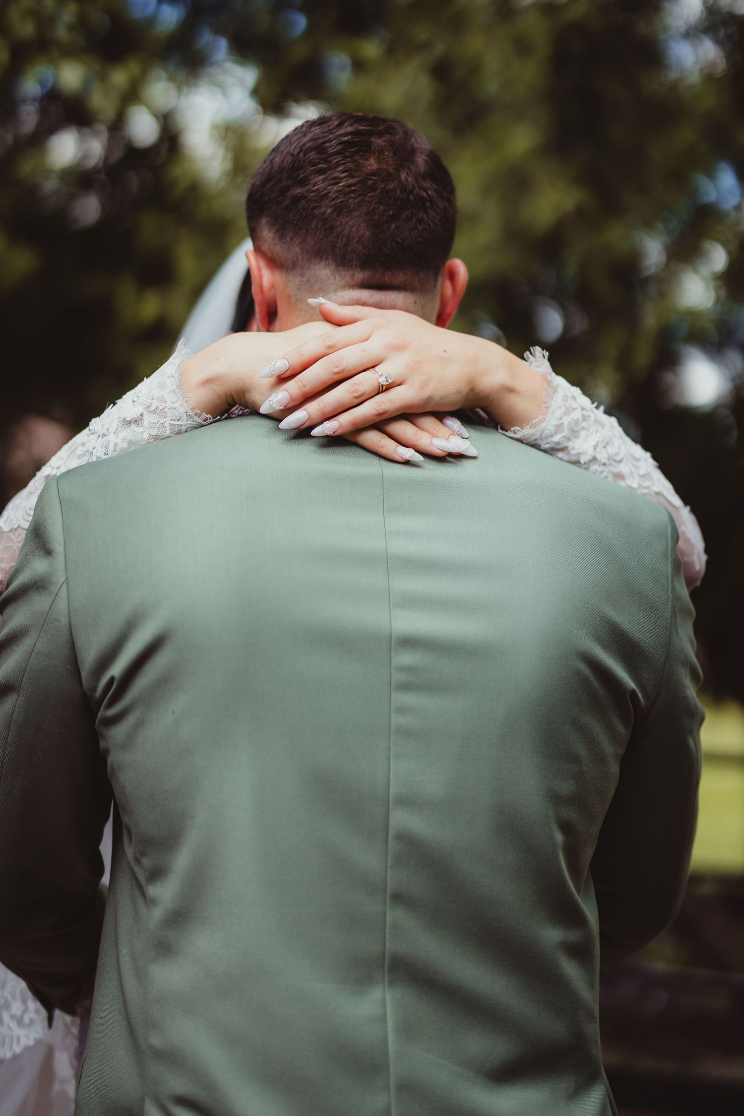Bride places her hand with wedding ring on grooms sage green suit jacket during outdoor wedding ceremony.