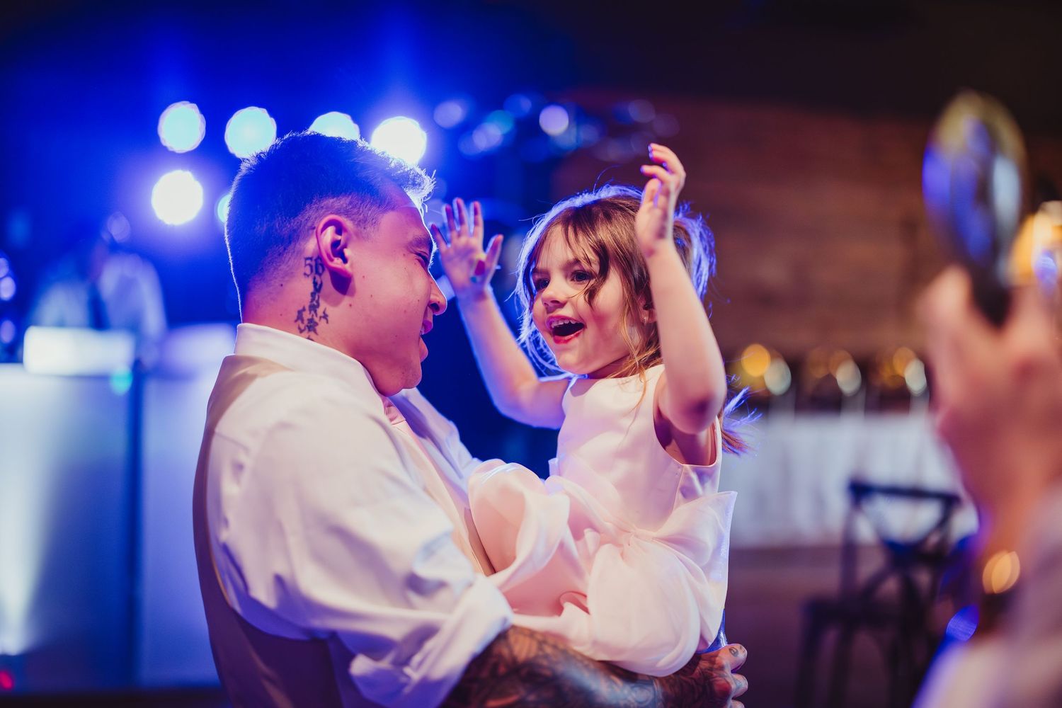 Adult holding a joyful child dancing at a wedding reception with blue stage lighting in background.