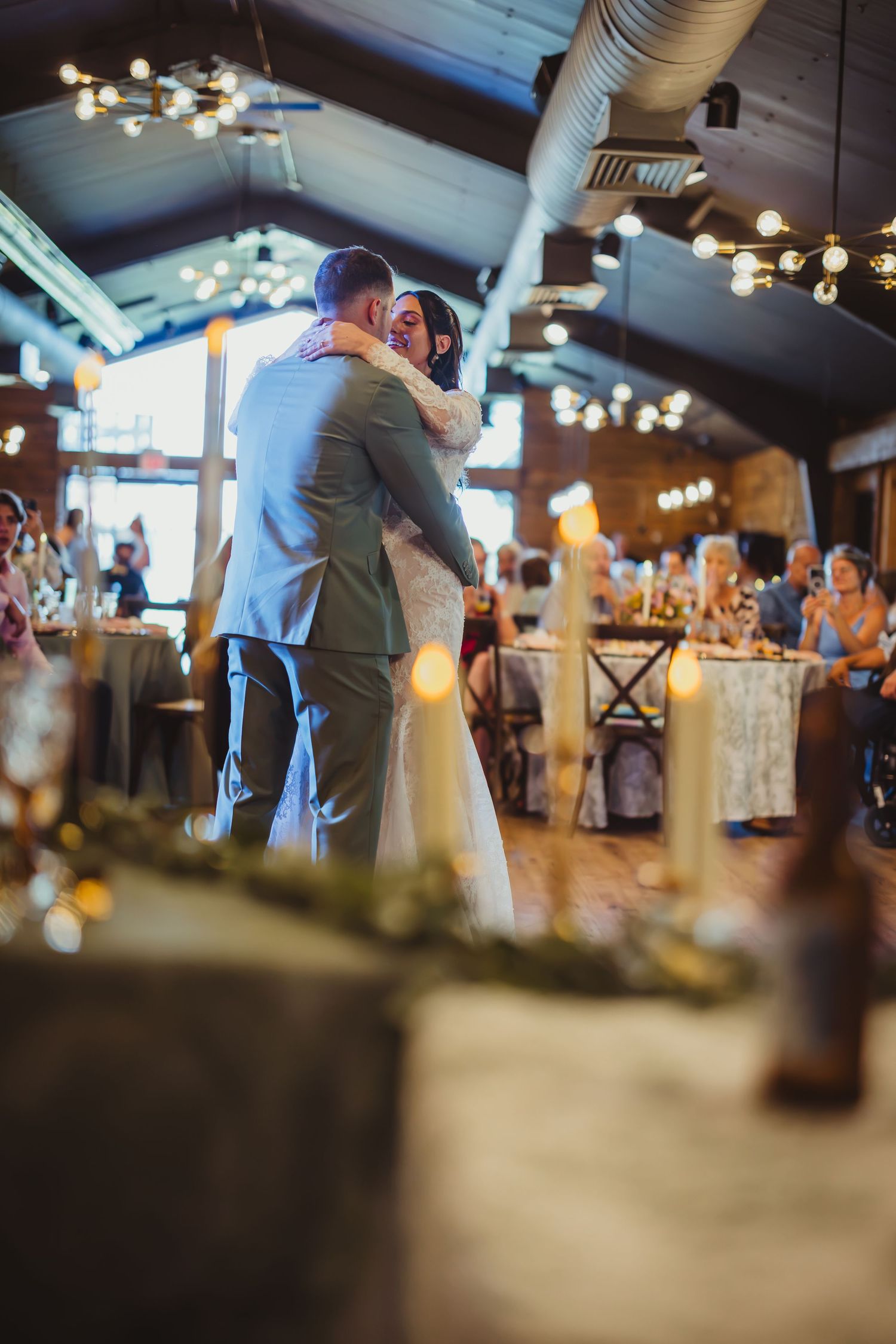 Bride and groom share their first dance in a warmly lit rustic wedding venue with string lights and candles.