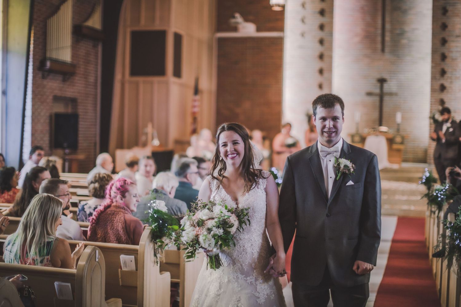 Church wedding celebration with guests watching ceremony from wooden pews lined with floral decorations.
