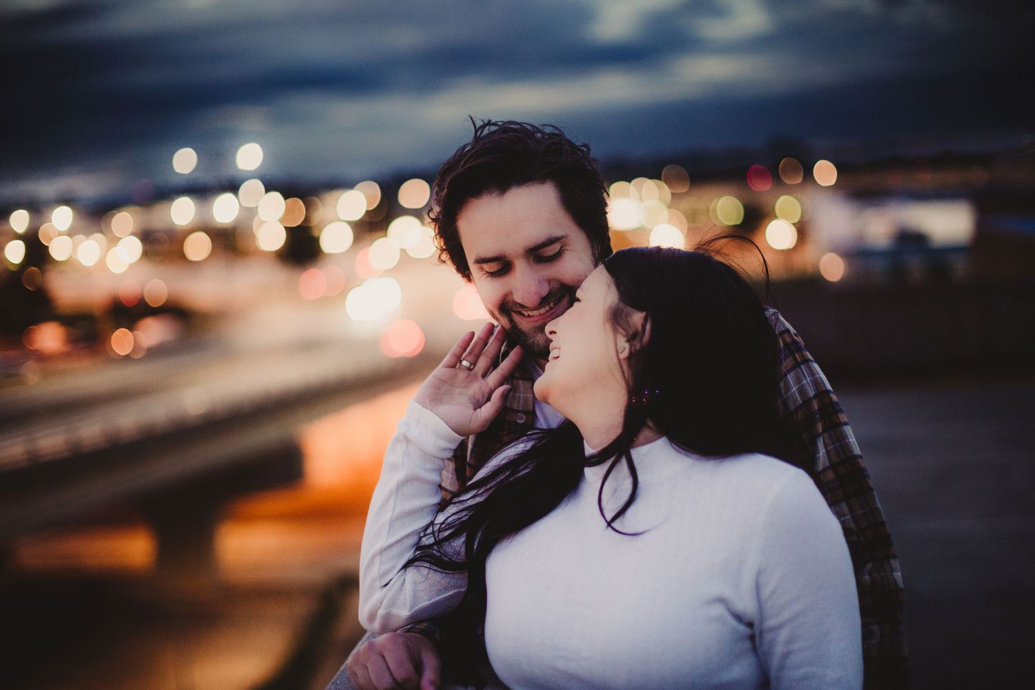 A couple shares an intimate moment against a blurred cityscape with bokeh lights at dusk.