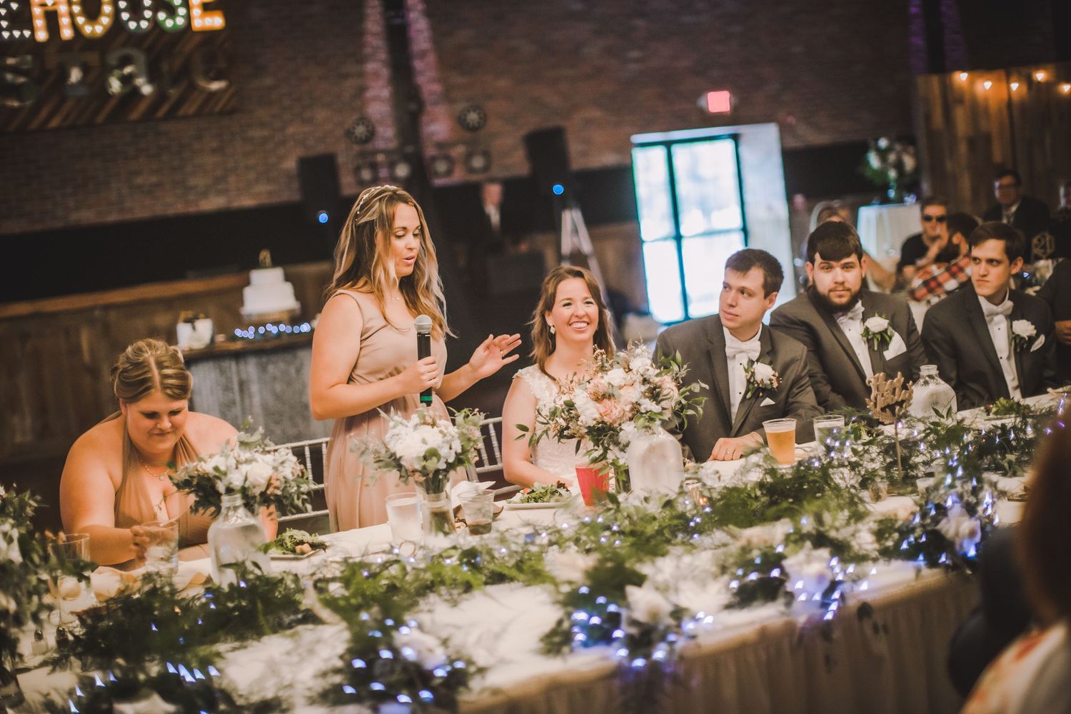 Wedding party members seated at a decorated head table giving toasts during the reception.